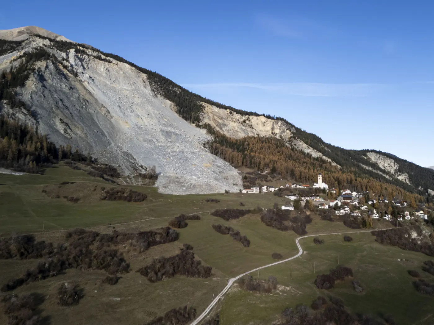 Una frana sulla montagna che sovrasta un villaggio di montagna con alberi di conifere, circondato da un paesaggio verde. Sulla destra, spicca un campanile bianco. Il cielo è sereno e blu.