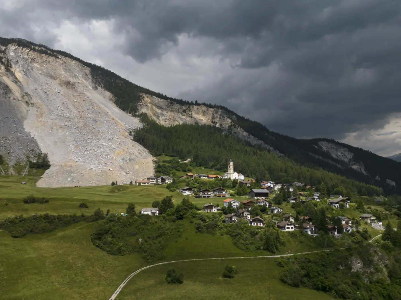 Un villaggio pittoresco si trova al piede di una montagna, con una chiesa visibile tra le case. Sullo sfondo, una grande frana di roccia si estende lungo la montagna, mentre nuvole scure coprono il cielo.