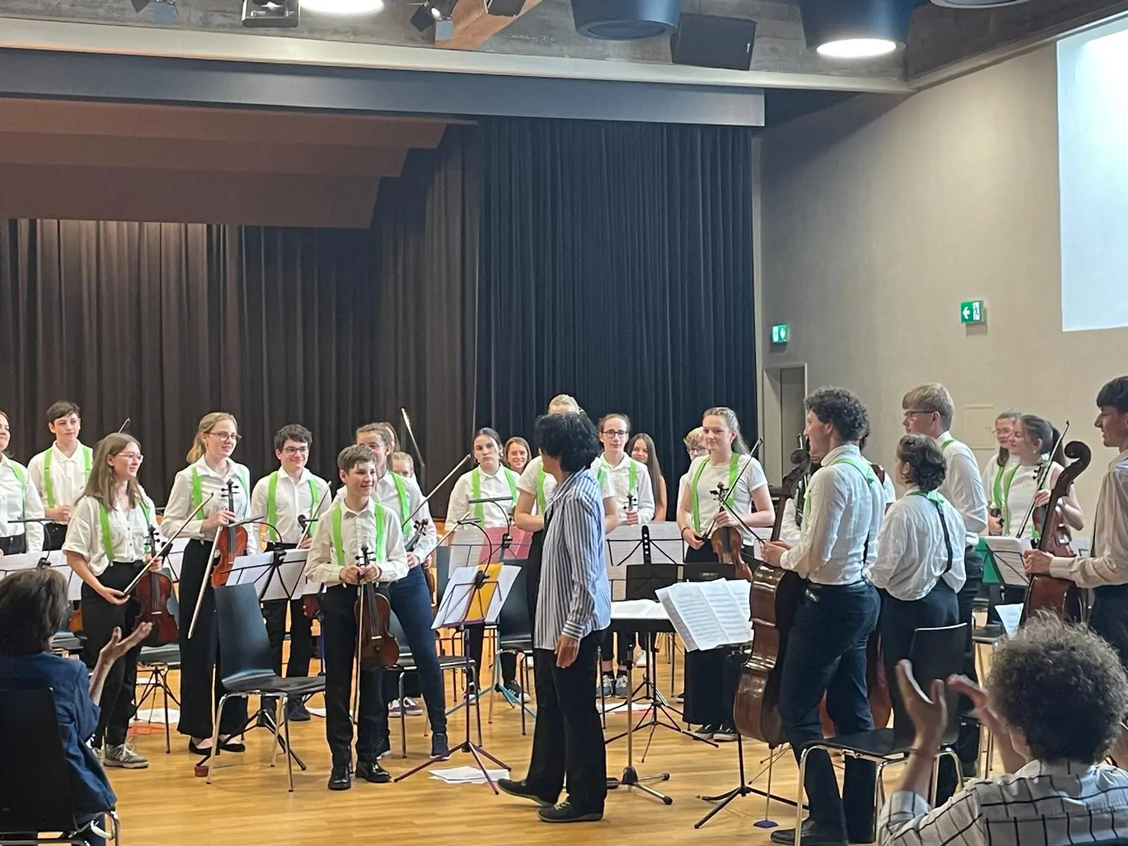 Un gruppo di giovani musicisti applauditi, vestiti di camicie bianche con cravatte verdi, si esibisce su un palco. Alcuni suonano strumenti orchestrali, mentre altri si preparano a ricevere riconoscimenti.