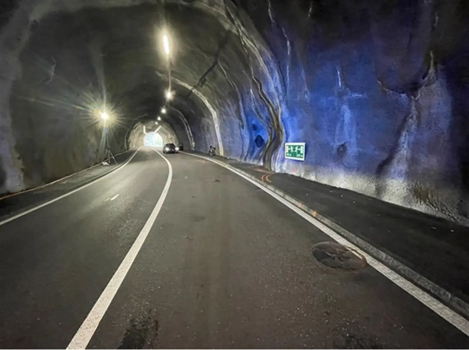 Tunnel con pareti rocciose illuminate da luci artificiali. La strada è parzialmente visibile e un'auto si avvicina all'uscita.