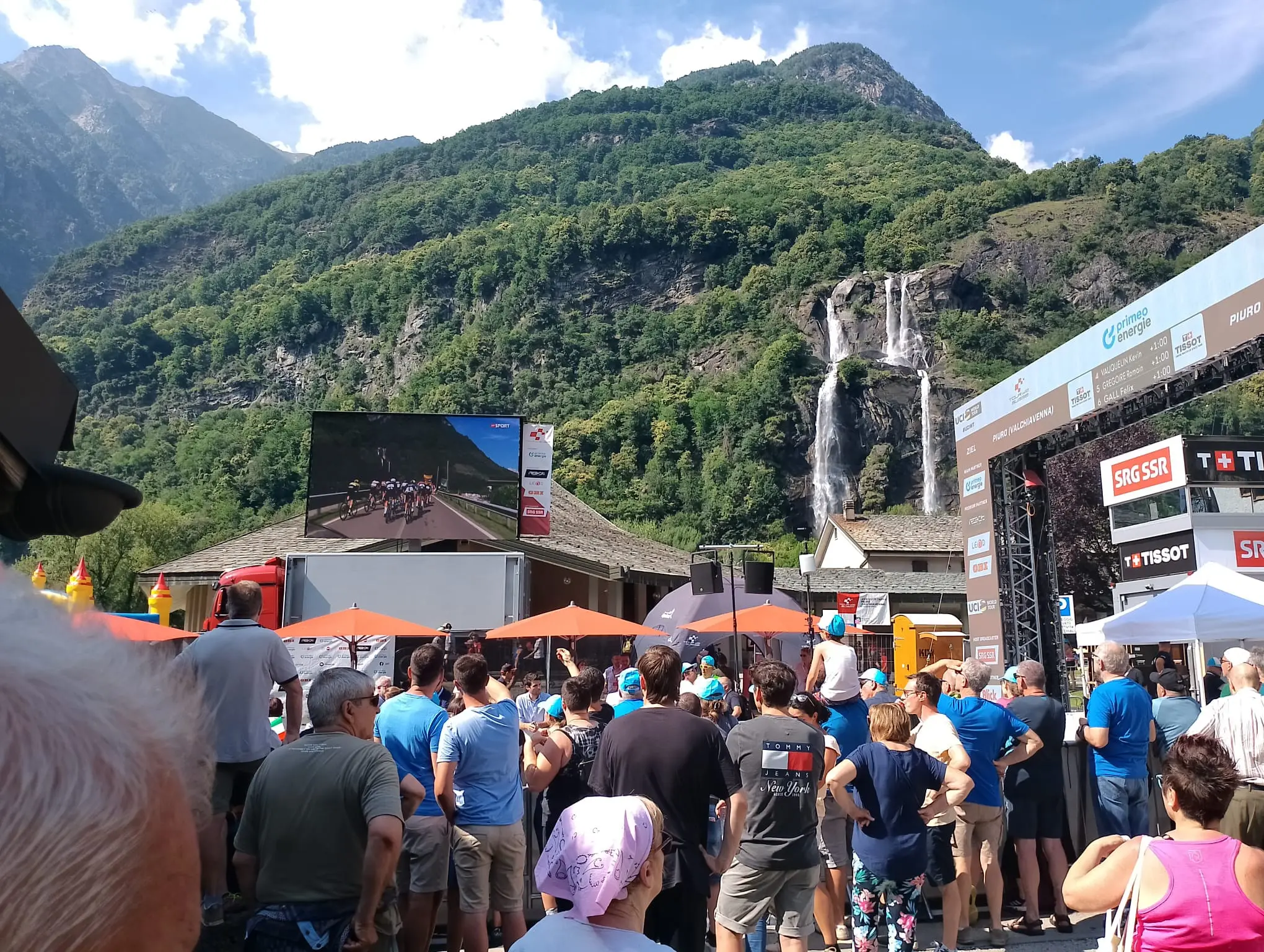 Una folla si raduna sotto un grande schermo che trasmette una gara di ciclismo. Sullo sfondo si vedono montagne verdi e una cascata. Personi vestiti in t-shirt azzurre e vari colori guardano con attenzione.