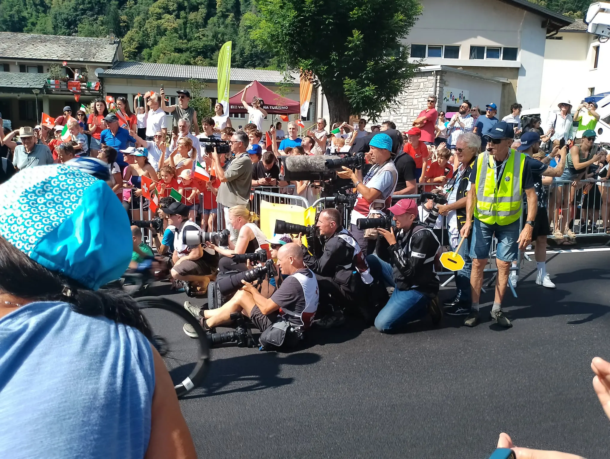 Un gruppo di fotografi e giornalisti accovacciati lungo una strada, circondati da una folla di spettatori. Alcuni indossano giubbotti luminosi e altri sono pronti con telecamere e attrezzature fotografiche. Sullo sfondo, si può vedere la vegetazione e alcuni tendoni.