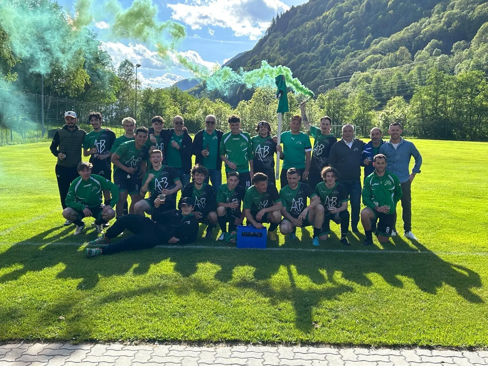 Squadra di calcio in uniforme verde, posando su un campo con il fondo montano. I giocatori sorridono e festeggiano, circondati da fumogeni verdi e blu.