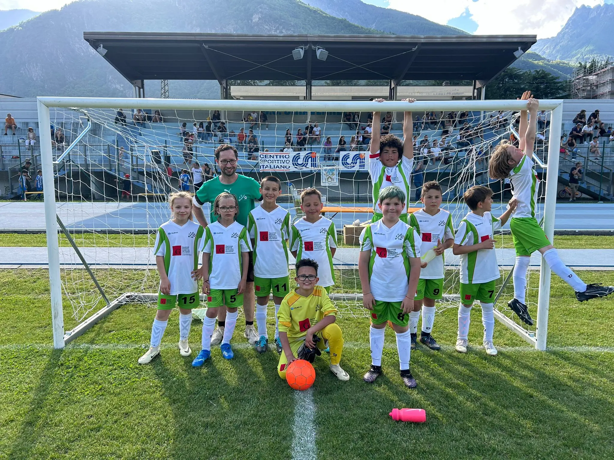 Una squadra di giovani calciatori sorride davanti a una porta da calcio. Indossano divise bianche e verdi. Alcuni si trovano in piedi, mentre uno è appeso alla rete. Sul campo verde, si vedono montagne sullo sfondo.