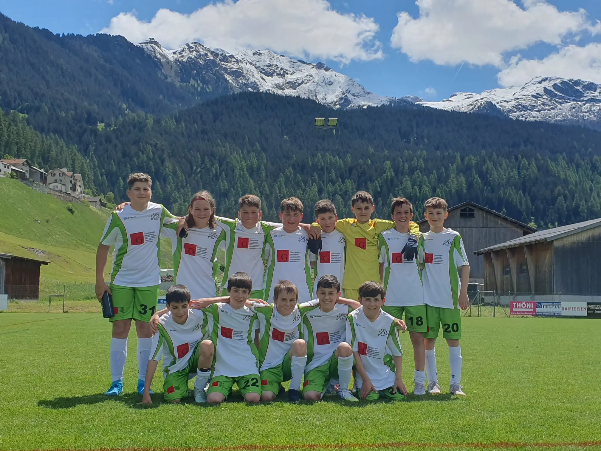 Una squadra di calcio giovanile in uniforme bianca e verde, sorridente e in posa, su un campo con montagne innevate sullo sfondo. Il cielo è sereno e soleggiato.