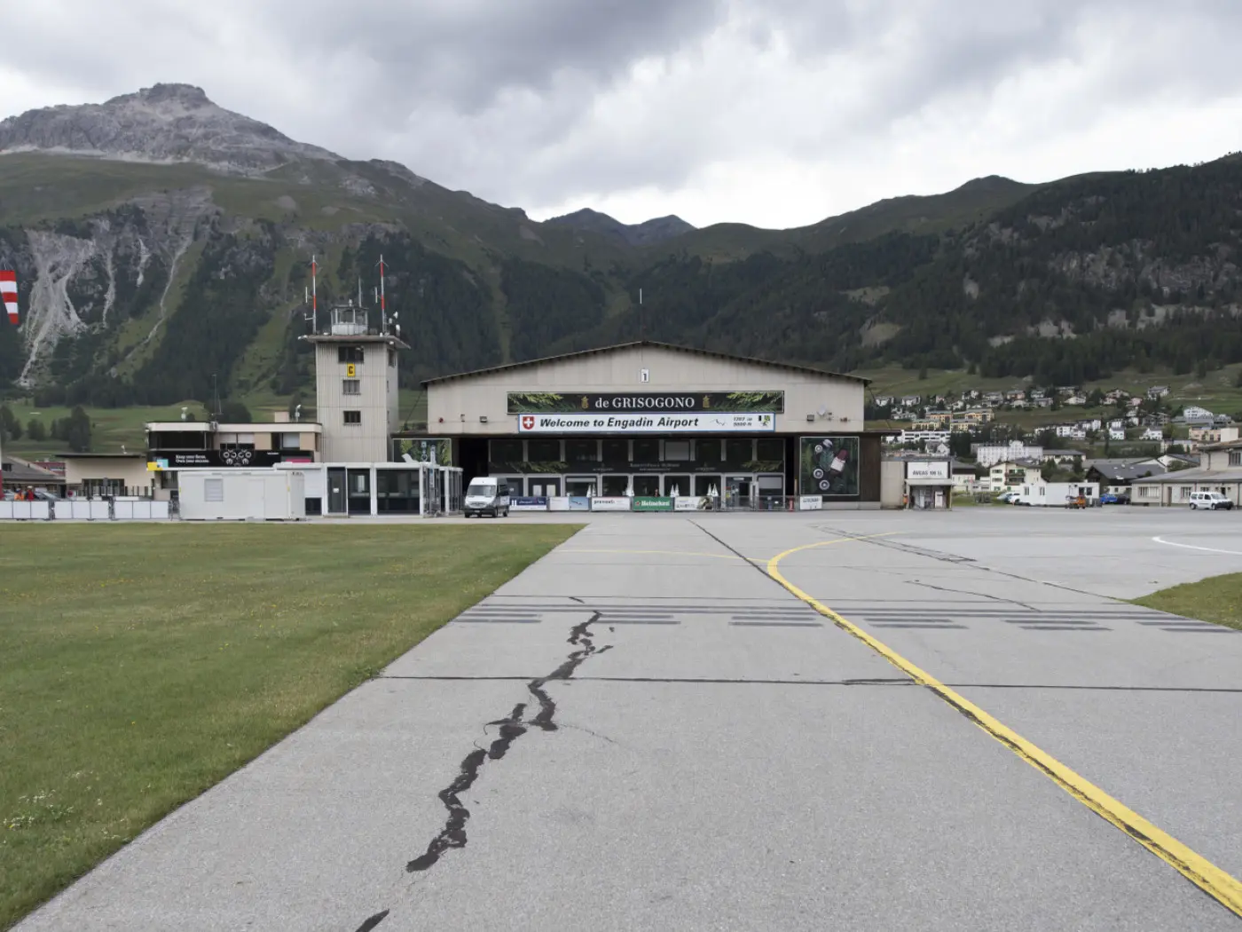 L'aeroporto di Samedan, visibile sotto un cielo nuvoloso, con montagne sullo sfondo e un'ampia pista in primo piano. La facciata dell'edificio principale mostra il nome dell'aeroporto e diverse bandiere.