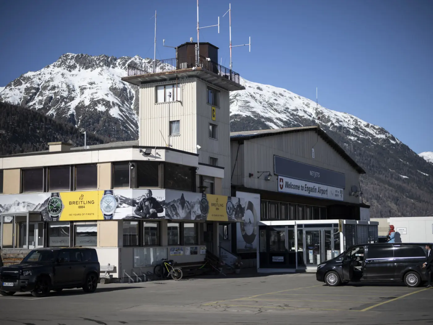 Edificio principale dell'aeroporto di Engadin, con un grande segnale di benvenuto e montagne innevate sullo sfondo. All'esterno, ci sono due veicoli parcheggiati.
