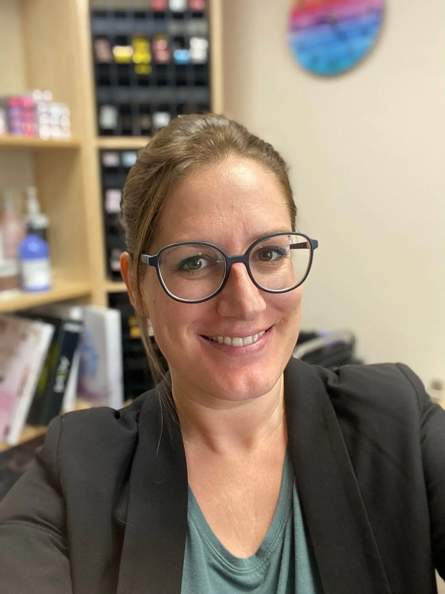 Donna sorridente con occhiali, capelli raccolti e giacca nera. Sullo sfondo, scaffali con prodotti e un quadro colorato.