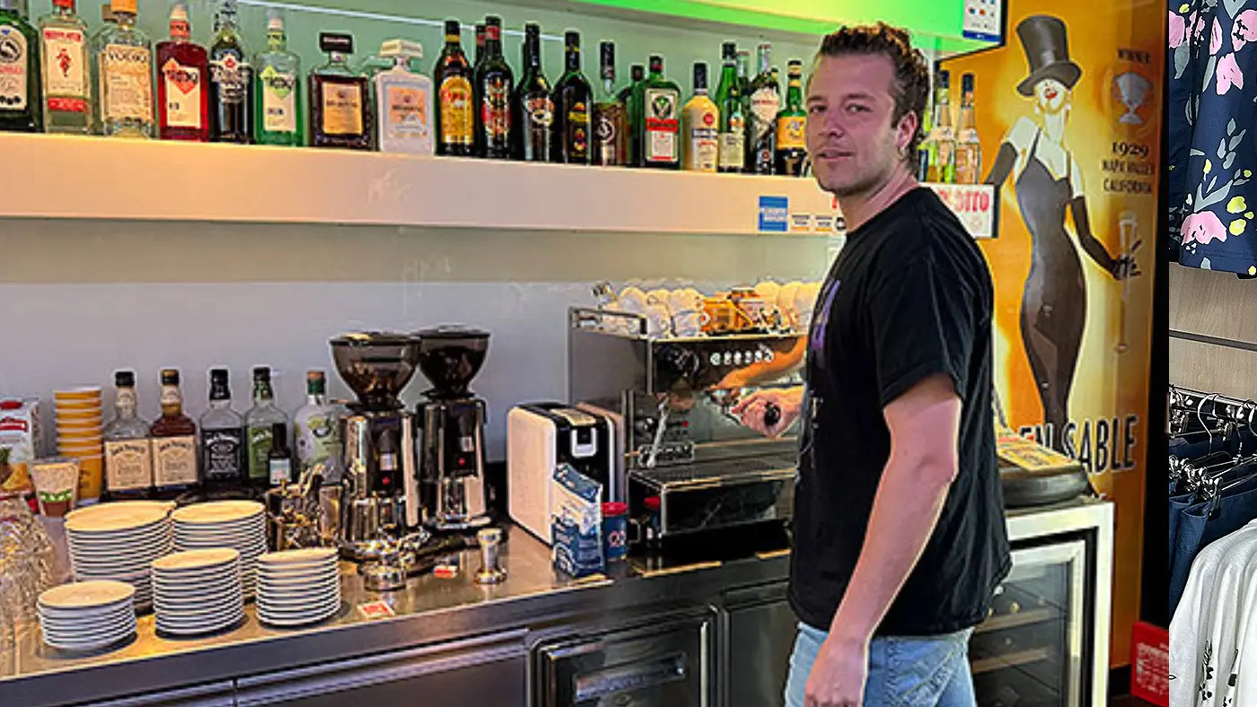 Un barista prepara bevande al bancone di un bar, con una macchina da caffè in funzione e scaffali pieni di bottiglie e bicchieri sullo sfondo.