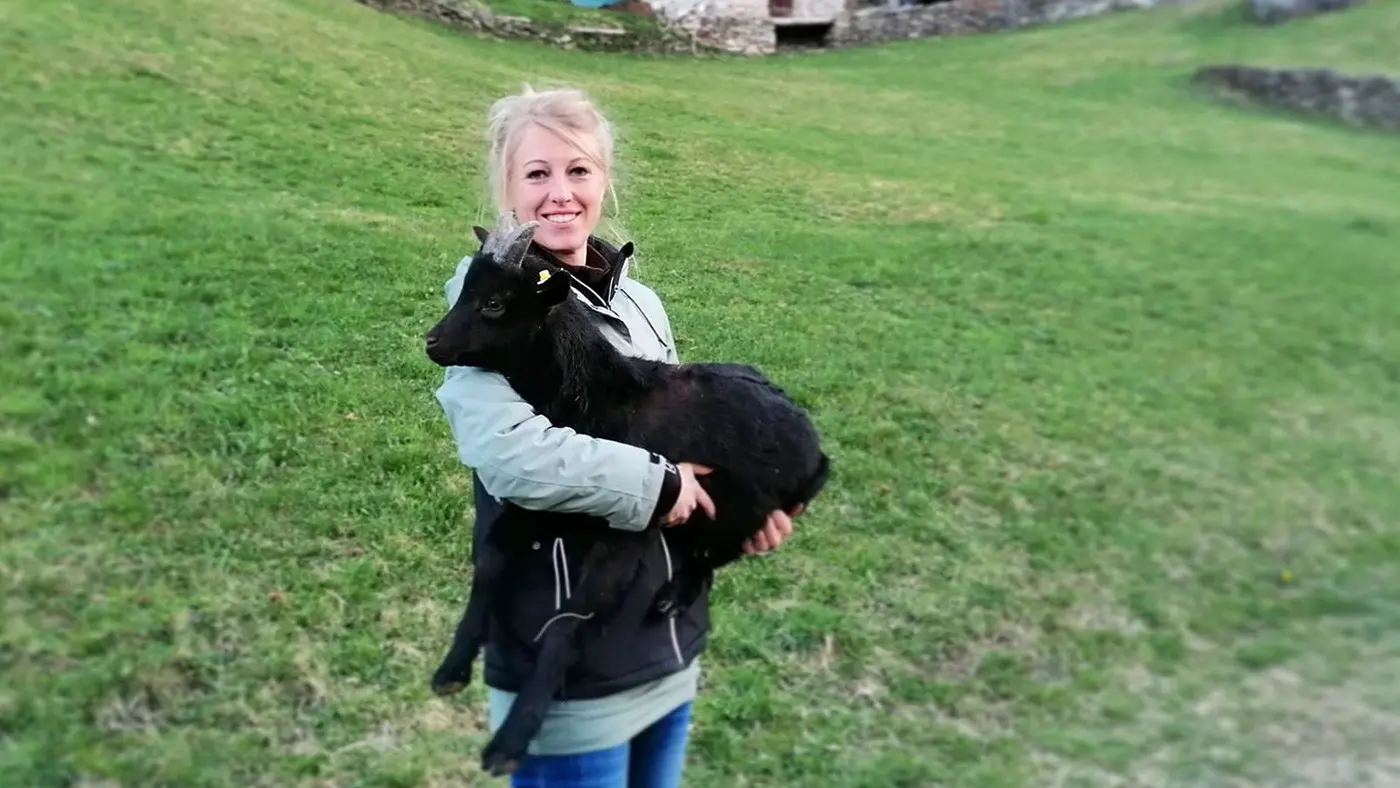 Una giovane donna sorride mentre tiene in braccio una capra nera su un prato verde. Sullo sfondo si vedono alcune strutture in pietra.