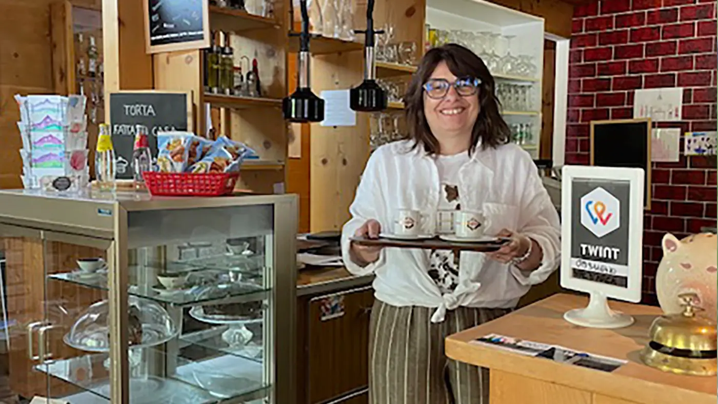Una donna sorridente tiene un vassoio con due tazzine di caffè, circondata da un ambiente di caffetteria rustico. Sullo sfondo ci sono dolci e un banco di servizio ben organizzato.