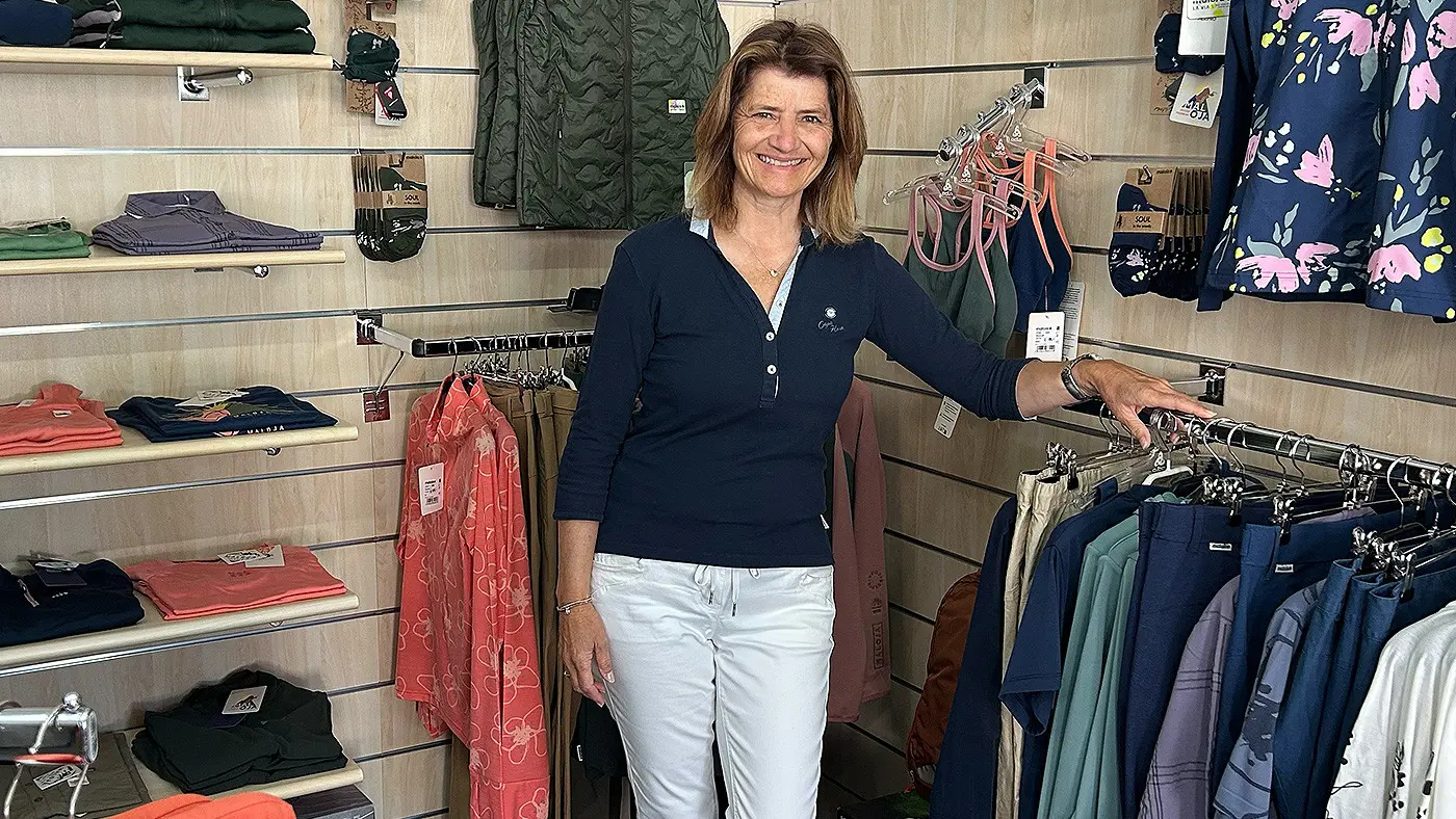 Donna sorridente in un negozio di abbigliamento, circondata da vari indumenti appesi. Indossa una maglia scura e pantaloni bianchi, con una mano appoggiata su una gruccia.