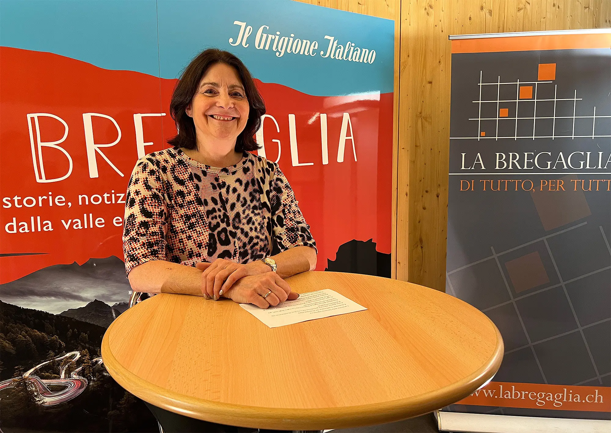Una donna sorride mentre posa dietro un tavolo rotondo, con un background che presenta un poster promozionale sulla Bregaglia. Indossa una maglietta a figura leopardata e tiene un foglio di carta.
