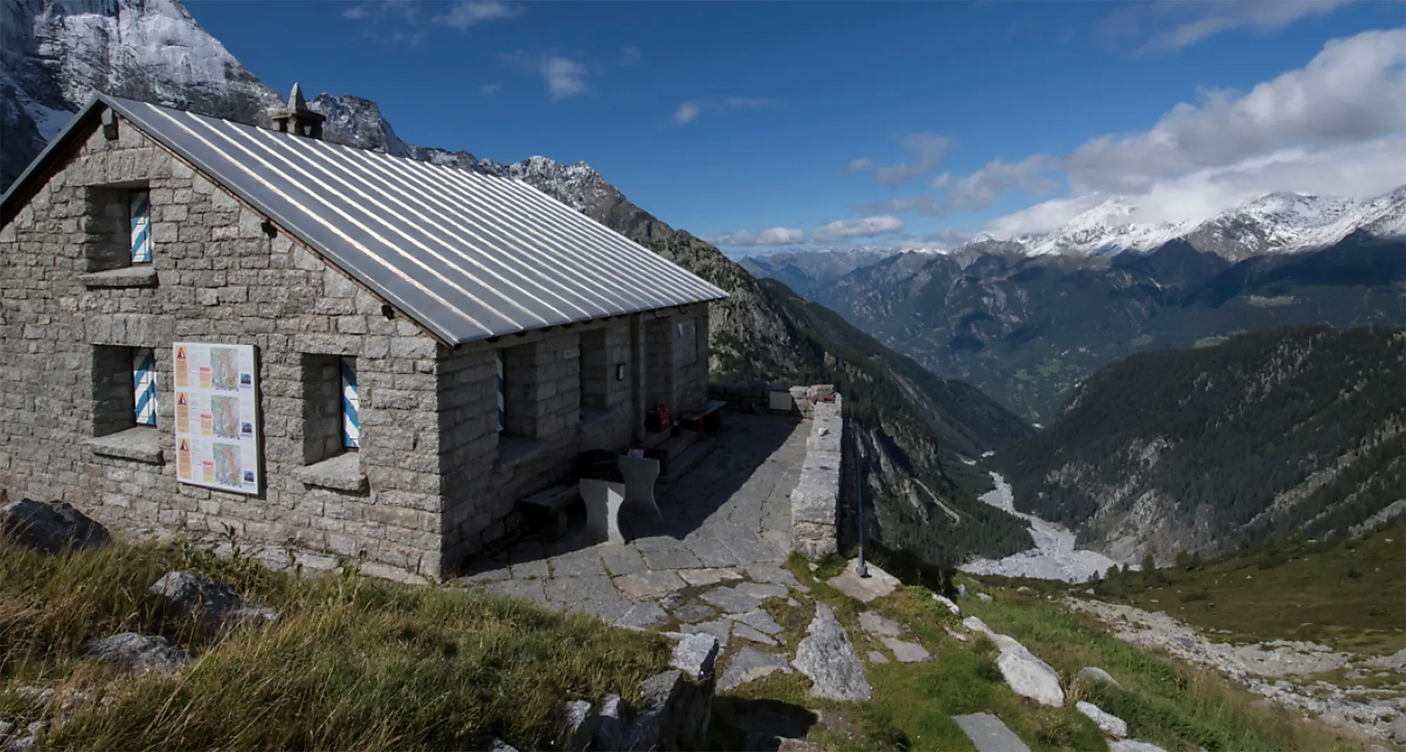 Una baita in pietra con un tetto di metallo, situata su una collina con vista sulle montagne. Il paesaggio circostante è verdeggiante, con valli e picchi innevati all'orizzonte. Cielo blu con alcune nuvole.