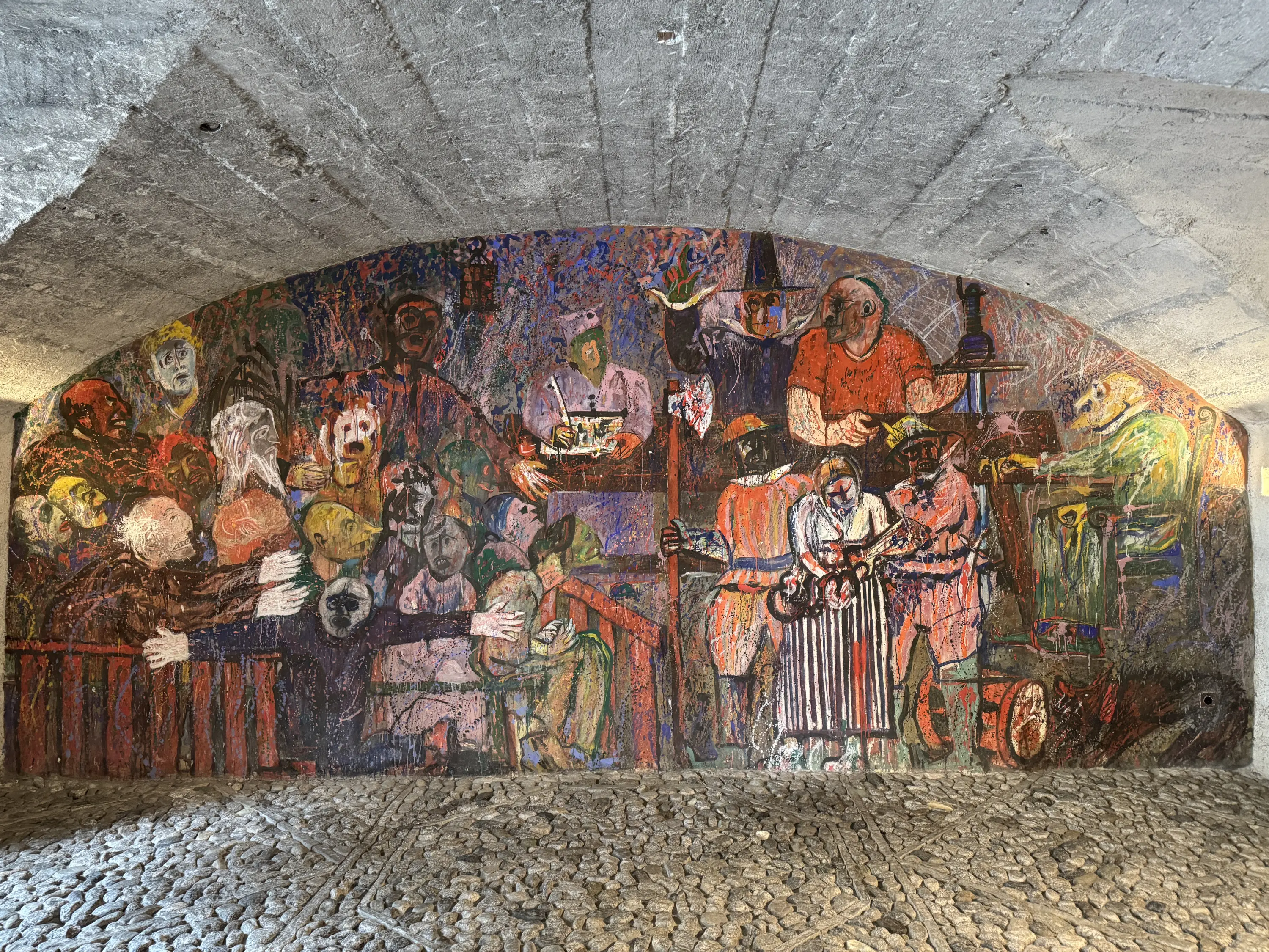 Murale colorato che rappresenta una scena di vita comunitaria, con figure umane che interagiscono tra loro. Le persone sono ritratte in diverse attività, creando un'atmosfera vivace e dinamica. Il fondo presenta colori intensi e tratti espressivi.