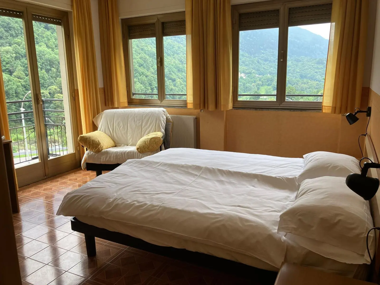 Camera luminosa con due finestre che affacciano su un paesaggio montano. C'è un letto matrimoniale con lenzuola bianche e un divano bianco con cuscini gialli. Le tende sono gialle e il pavimento è in piastrelle.