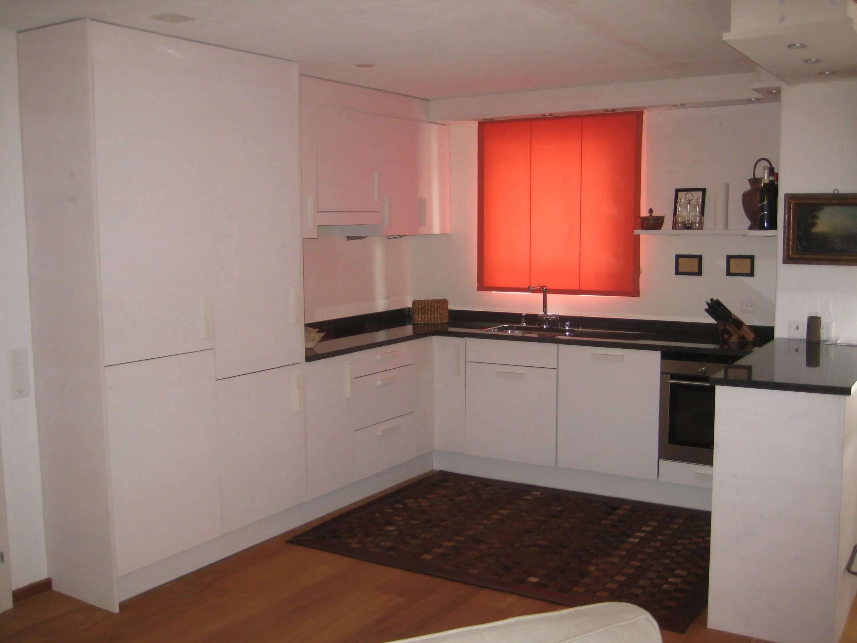 Cucina moderna con mobili bianchi e piano di lavoro in granito scuro. Una finestra con tendina rossa illumina l'ambiente. Un tappeto marrone è posizionato sul pavimento in legno.