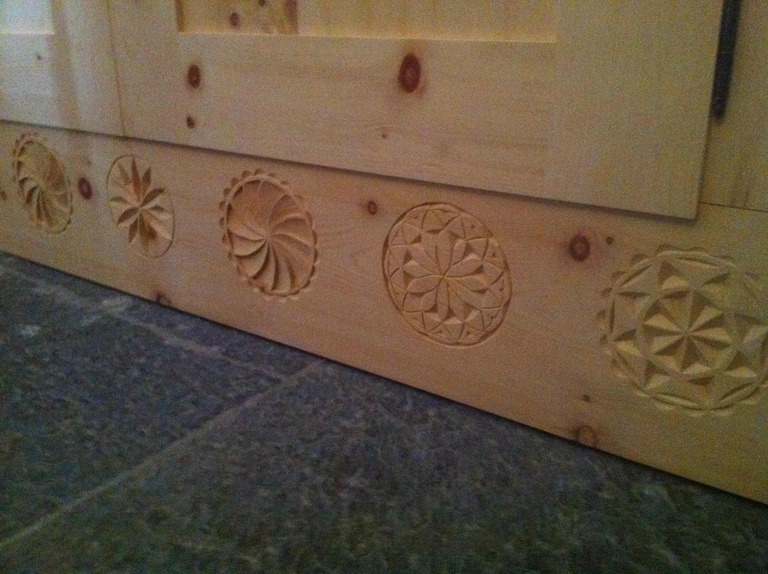 Rivestimento in legno con intagli decorativi. Vi sono cinque disegni circolari incisi, ciascuno con motivi geometrici differenti. Il legno è chiaro e il pavimento è di pietra grigia.