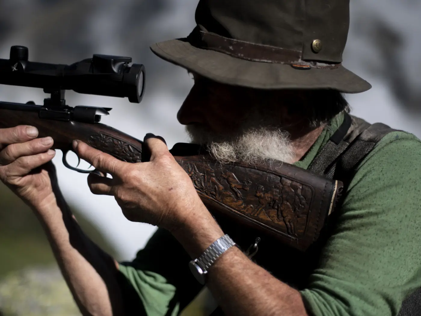 Uomo anziano con barba grigia che mira con un fucile, indossando un cappello e una camicia verde. Il fucile presenta decorazioni elaborate e il fondo mostra un paesaggio montano sfocato.