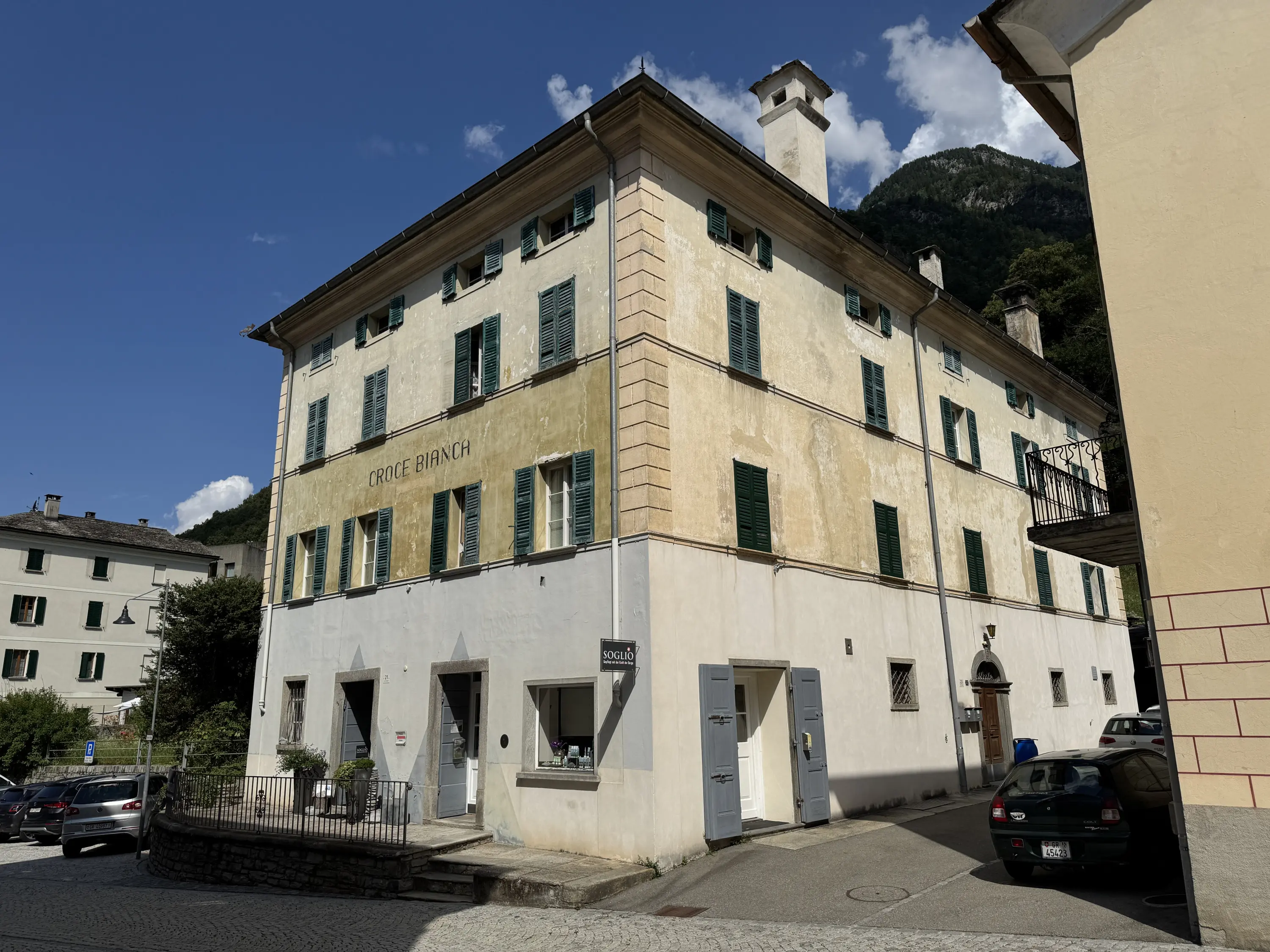 Edificio storico con facciata gialla e finestre verdi, situato in una piazza con montagne sullo sfondo. La scritta "CROICE BRANCH" è visibile sulla parete. Automobili parcheggiate davanti e un'area di giardino curata.