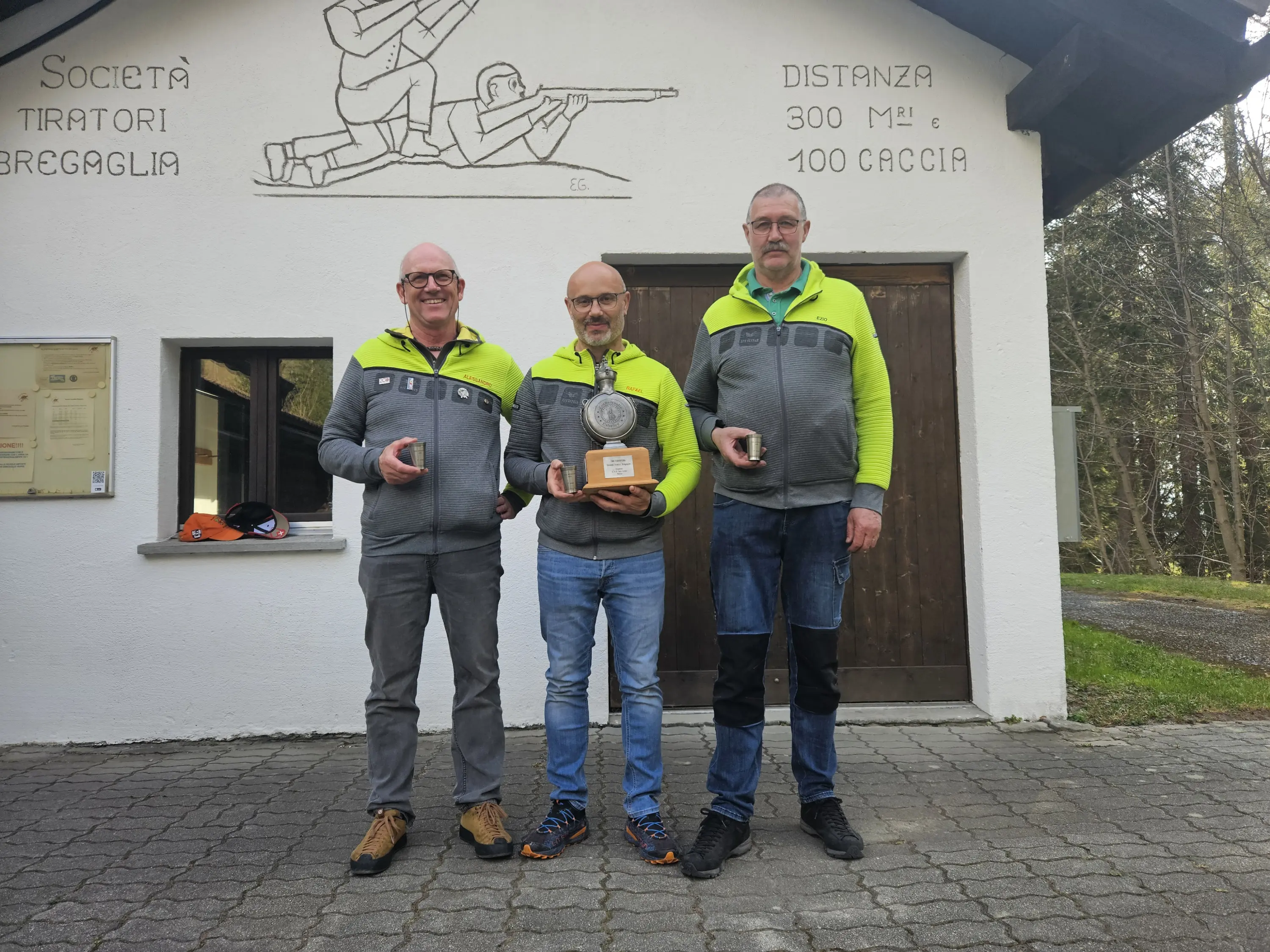 Tre uomini sorridenti sono in piedi davanti a un edificio con una targa sopra la porta. Indossano giacche verde fluorescente e tengono in mano dei trofei.