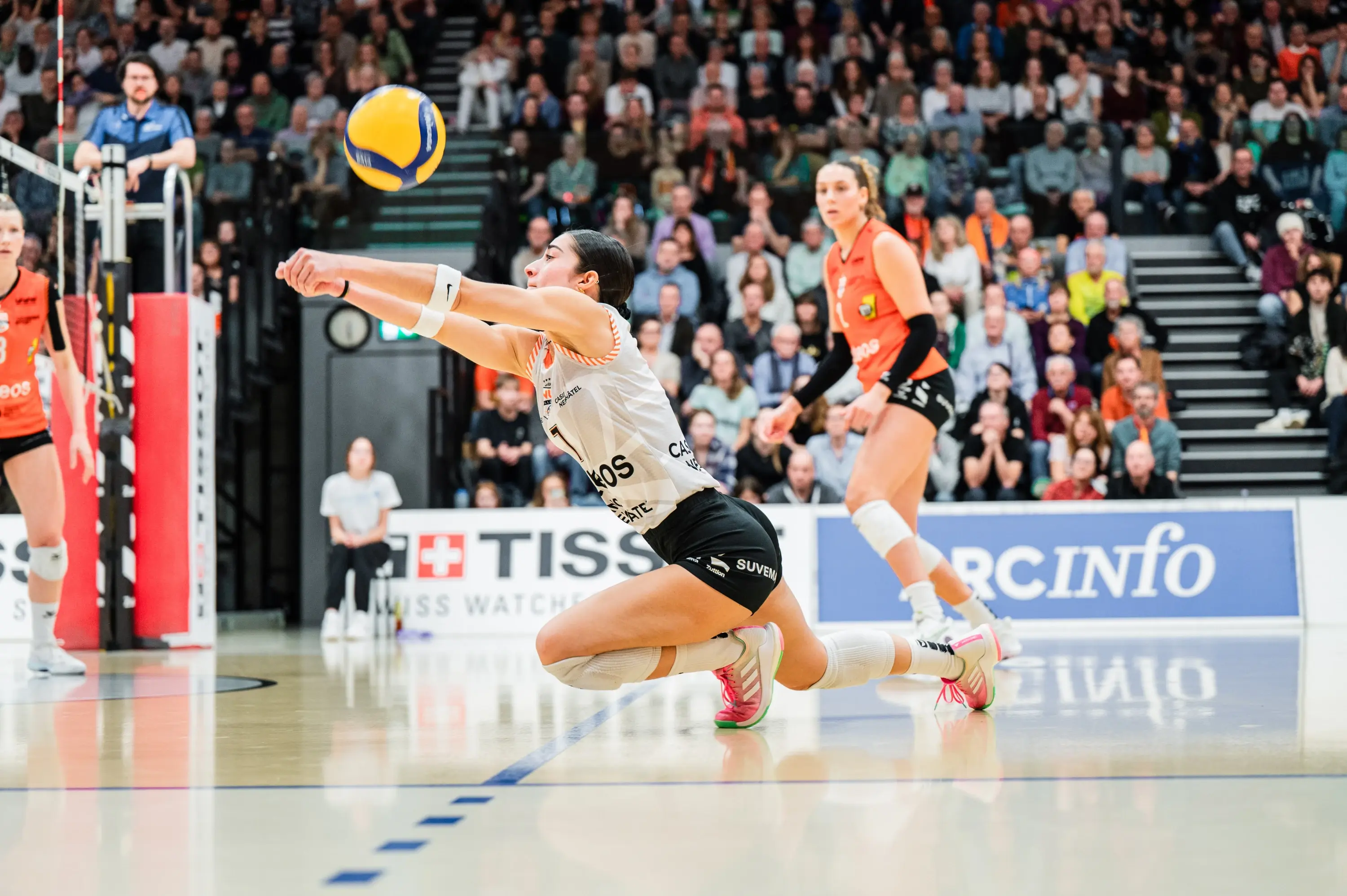 Giocatrice di pallavolo in tuffo per ricevere un pallone durante una partita, con un pubblico numeroso sullo sfondo. Altre giocatrici sono visibili sul campo.