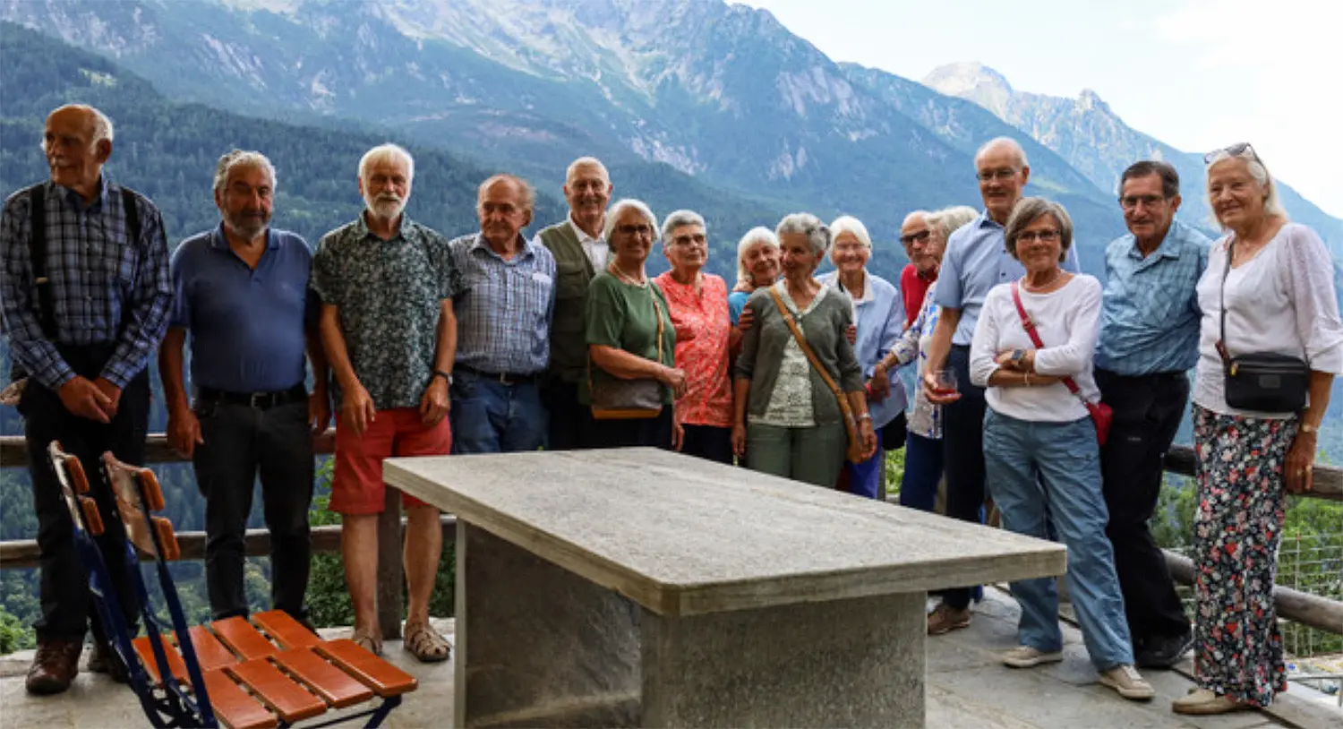 Un gruppo di quindici persone in posa davanti a una tavola di pietra, con montagne e natura sullo sfondo. Alcuni sorridono mentre altri sembrano più seri. Indossano abbigliamento estivo e vari colori.