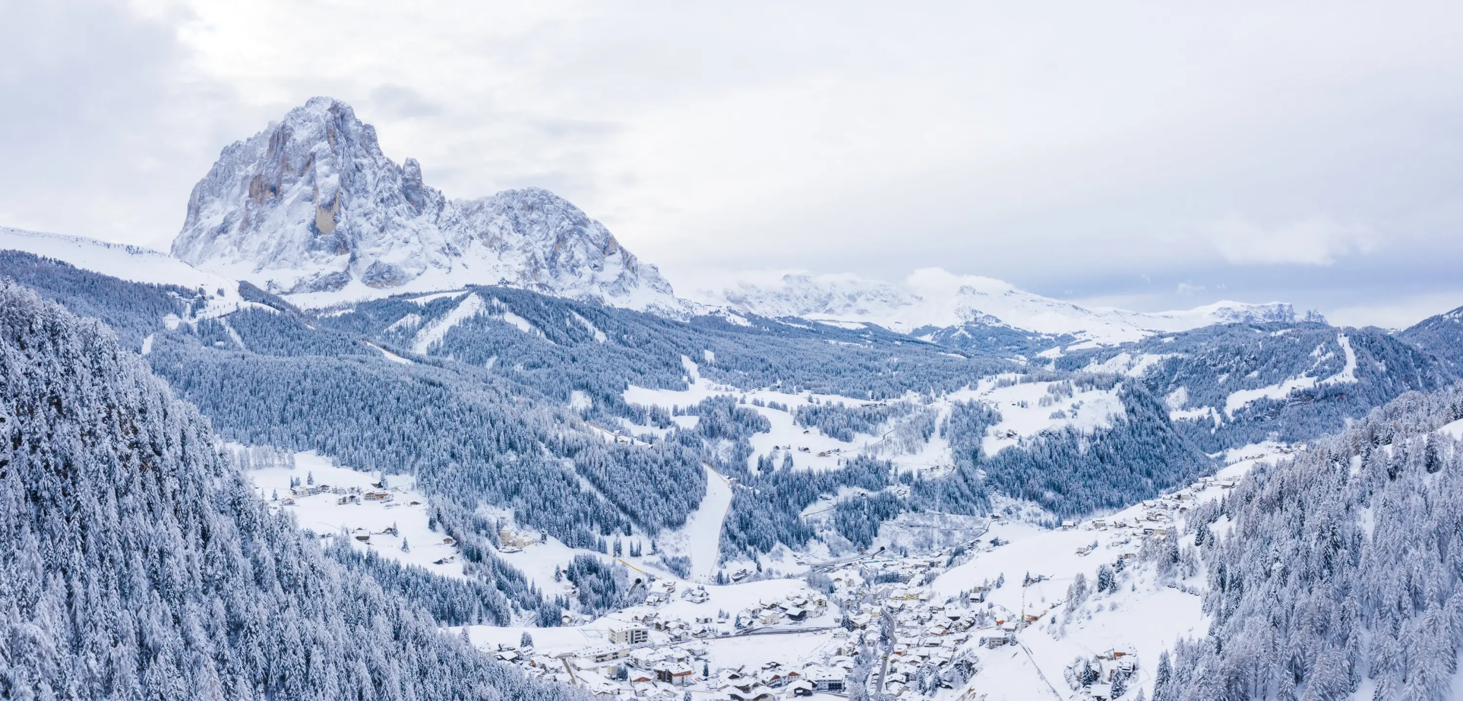 Panorama montano con un vasto paesaggio innevato. La cima di una montagna prominente si staglia all'orizzonte, circondata da foreste di conifere coperte di neve. Un villaggio alpino è visibile in basso, con case sparse tra le colline innevate. Il cielo è grigio e nuvoloso, creando un'atmosfera invernale.