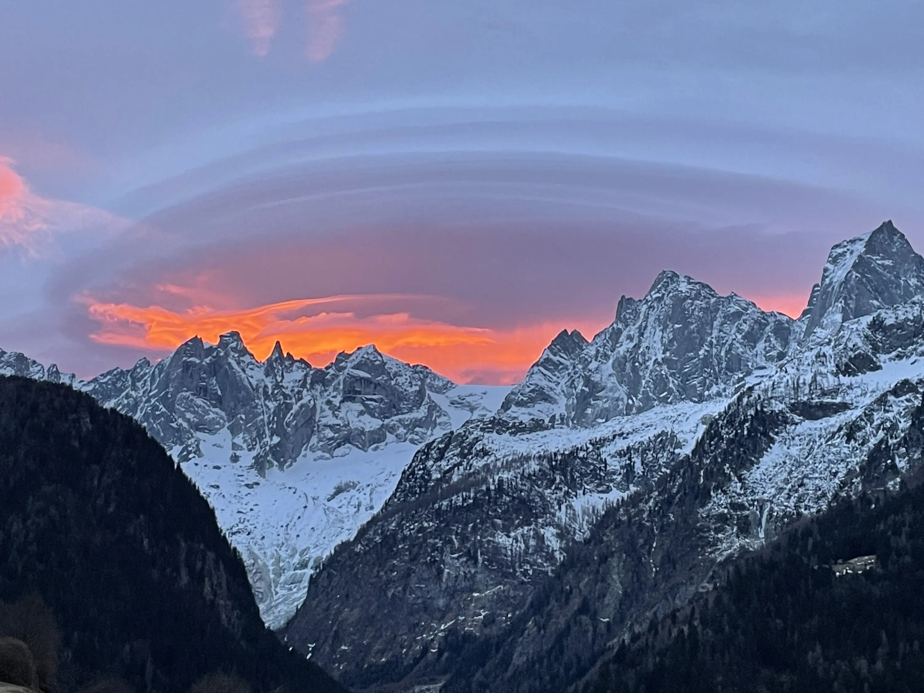 Il tramonto illumina le cime delle montagne innevate, creando un gioco di colori tra il rosa e l'arancione nel cielo, mentre nuvole a forma di anello si distribuiscono sopra.