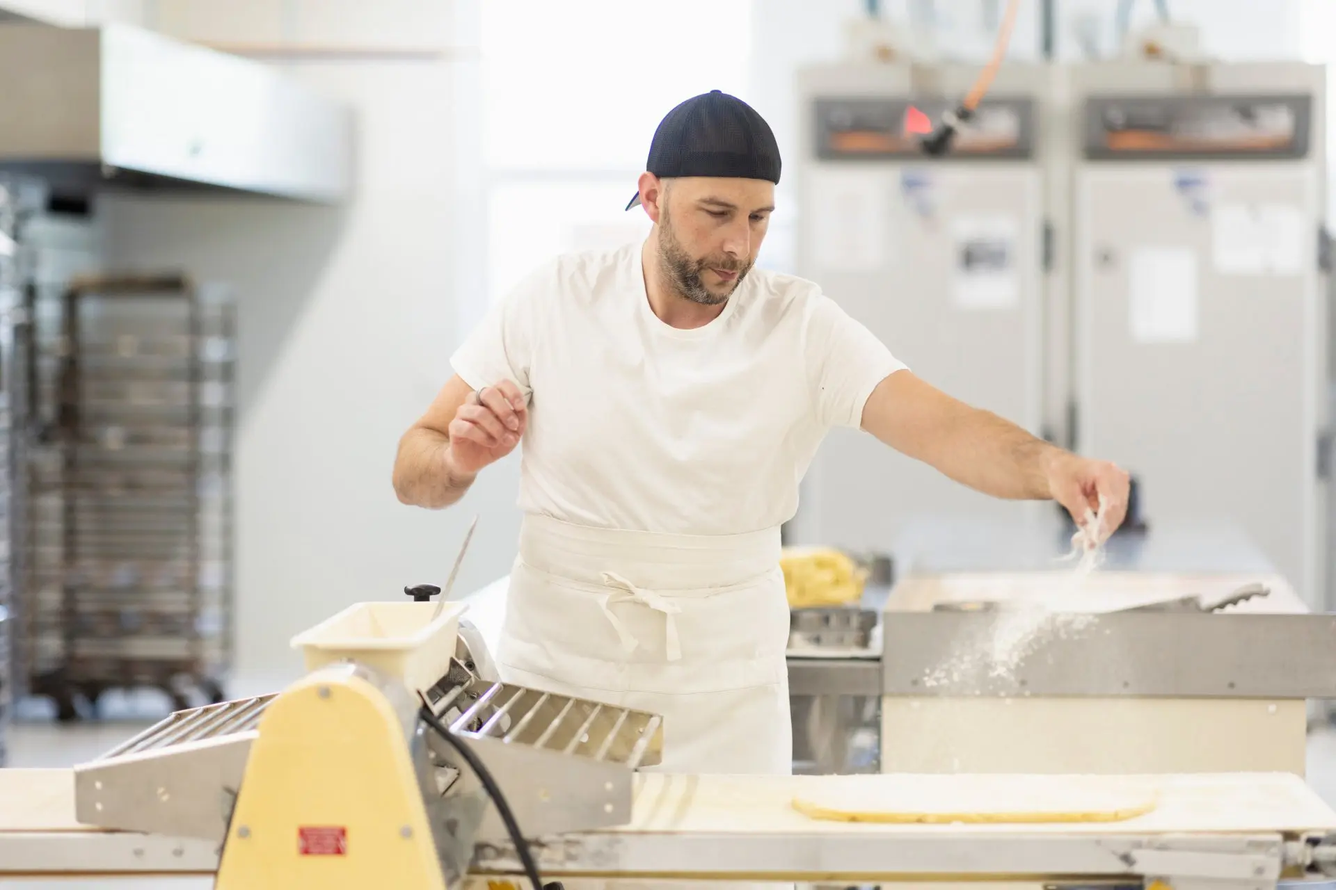 Un panettiere indossa un cappellino mentre spruzza farina su un impasto steso su un tavolo da lavoro. Sullo sfondo ci sono attrezzature professionali e ripiani con forni.