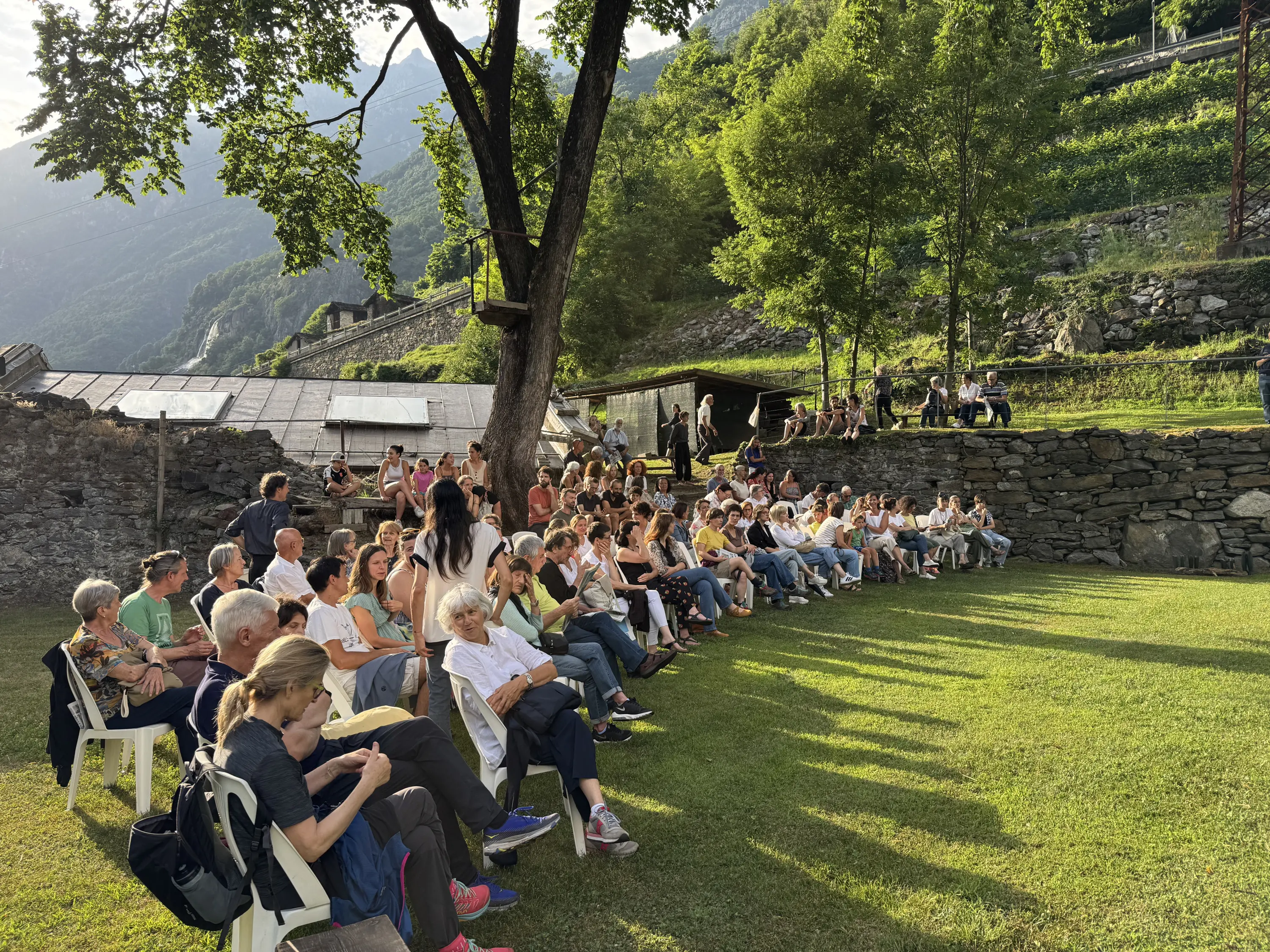 Un pubblico numeroso seduto su sedie bianche in un'area verde, circondato da alberi e colline. Sullo sfondo, si vedono edifici e strutture in pietra. La scena è illuminata dalla luce del sole al tramonto.