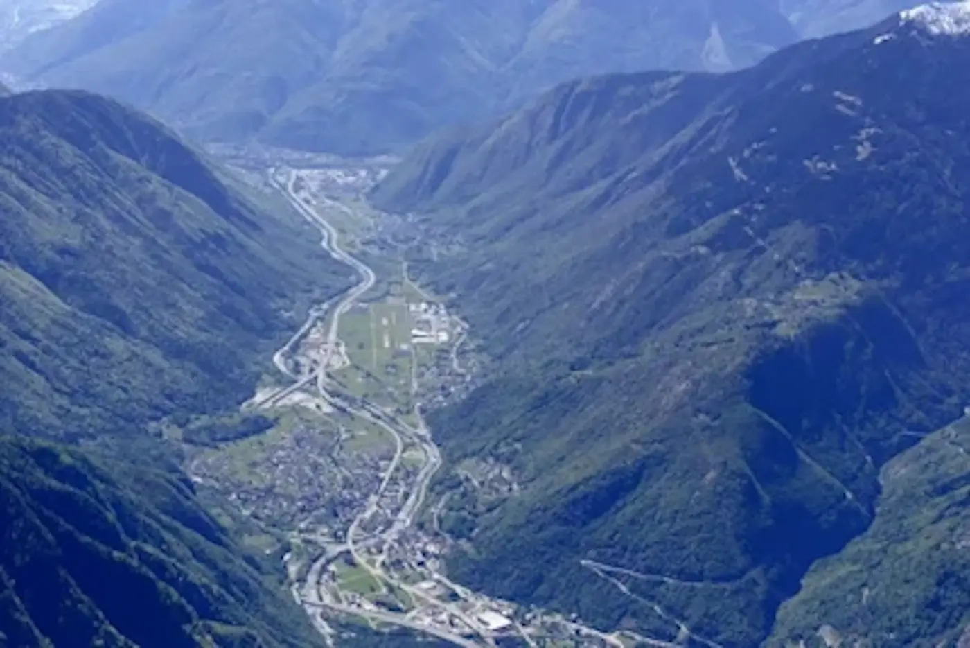 Una vista panoramica di una valle circondata da montagne, con un fiume e strade che serpeggiano tra il paesaggio. Un piccolo aeroporto è visibile in lontananza.