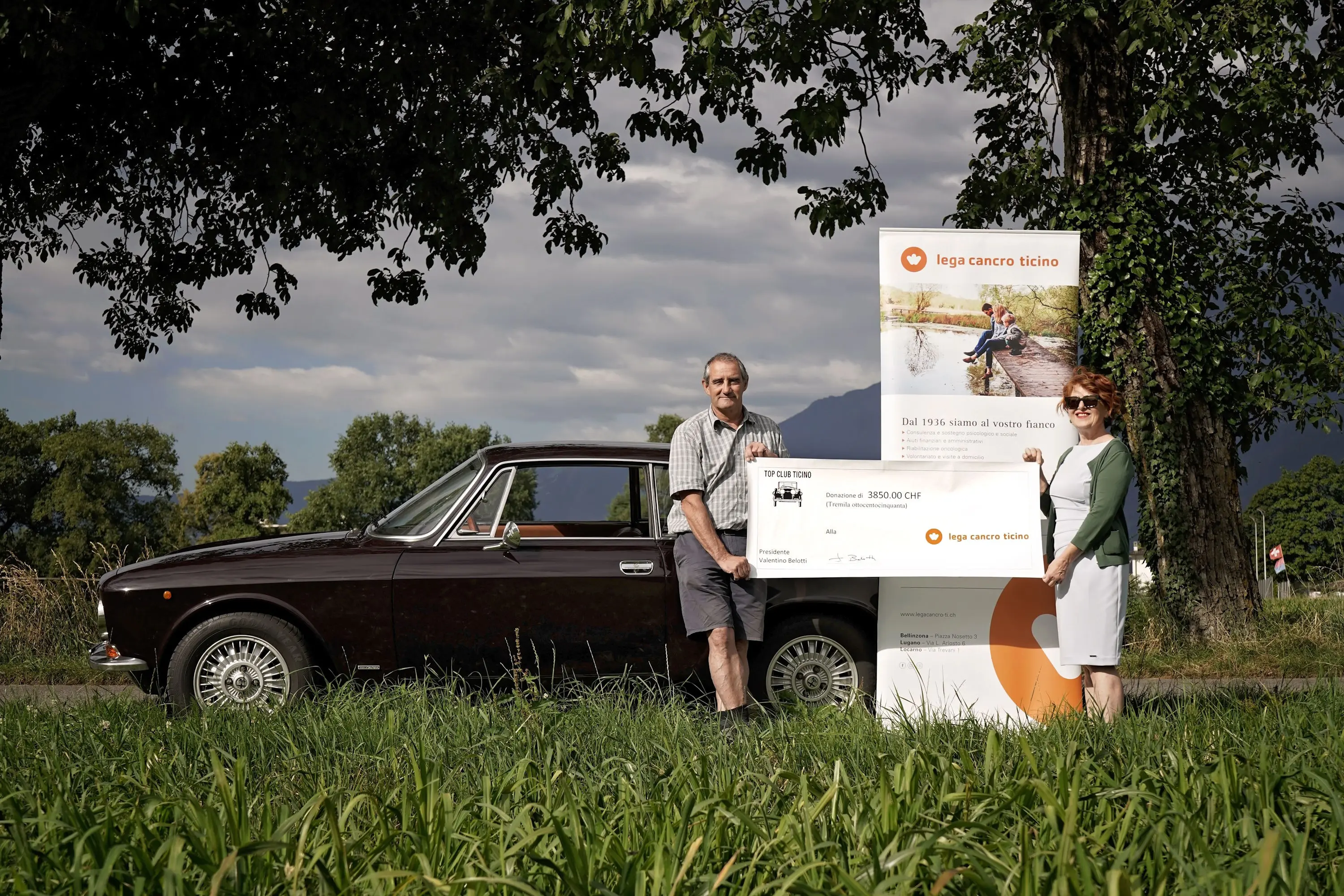 Un uomo e una donna sorridono accanto a un’auto d’epoca, mentre mostrano un grande assegno. Dietro di loro, un cartello della Lega contro il cancro del Ticino, con un paesaggio naturale sullo sfondo.