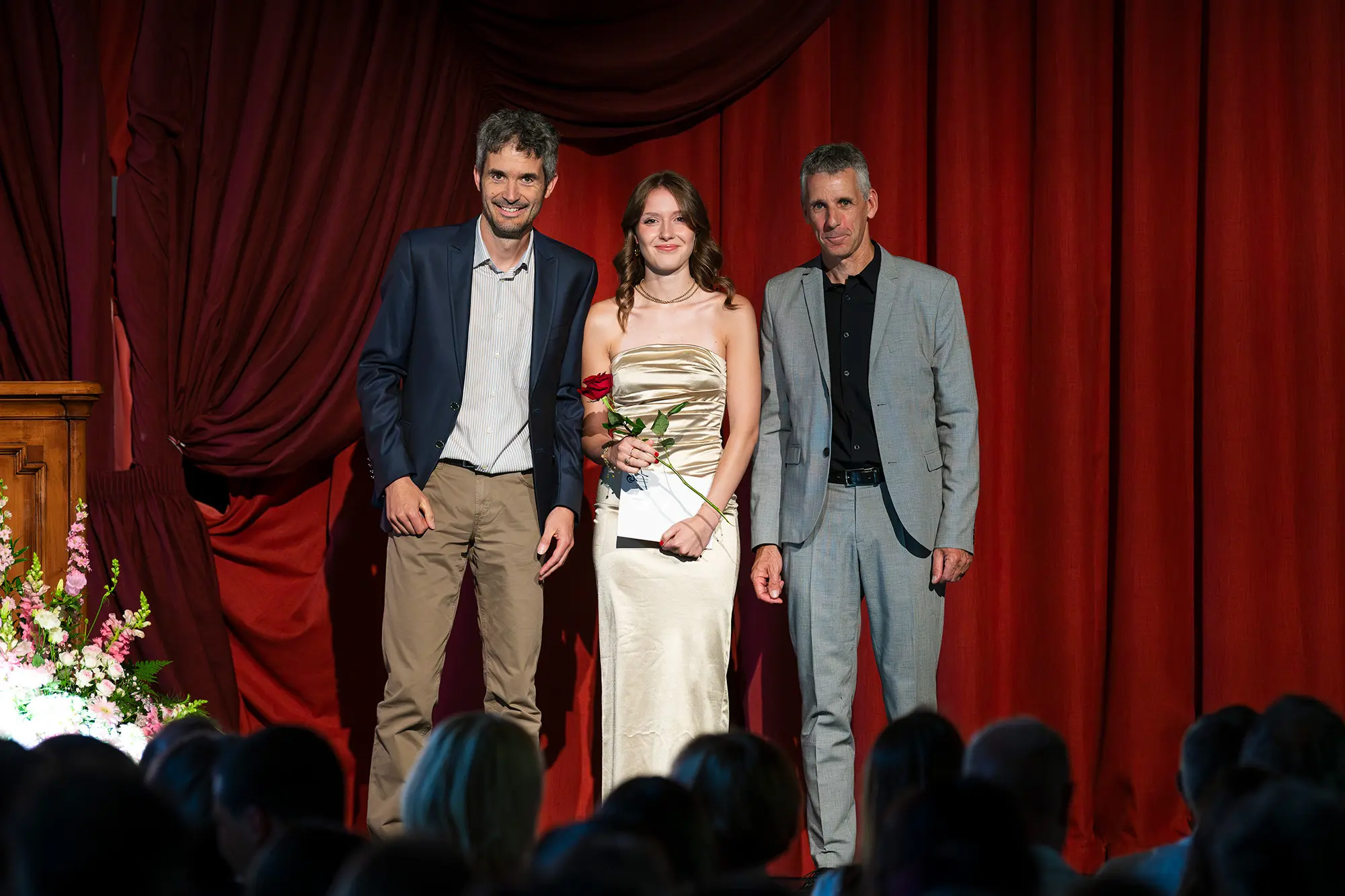 Tre persone sul palco: due uomini in abiti eleganti e una giovane donna in un abito dorato con una rosa in mano. Si trovano di fronte a una tenda rossa e un pubblico visibile in basso.