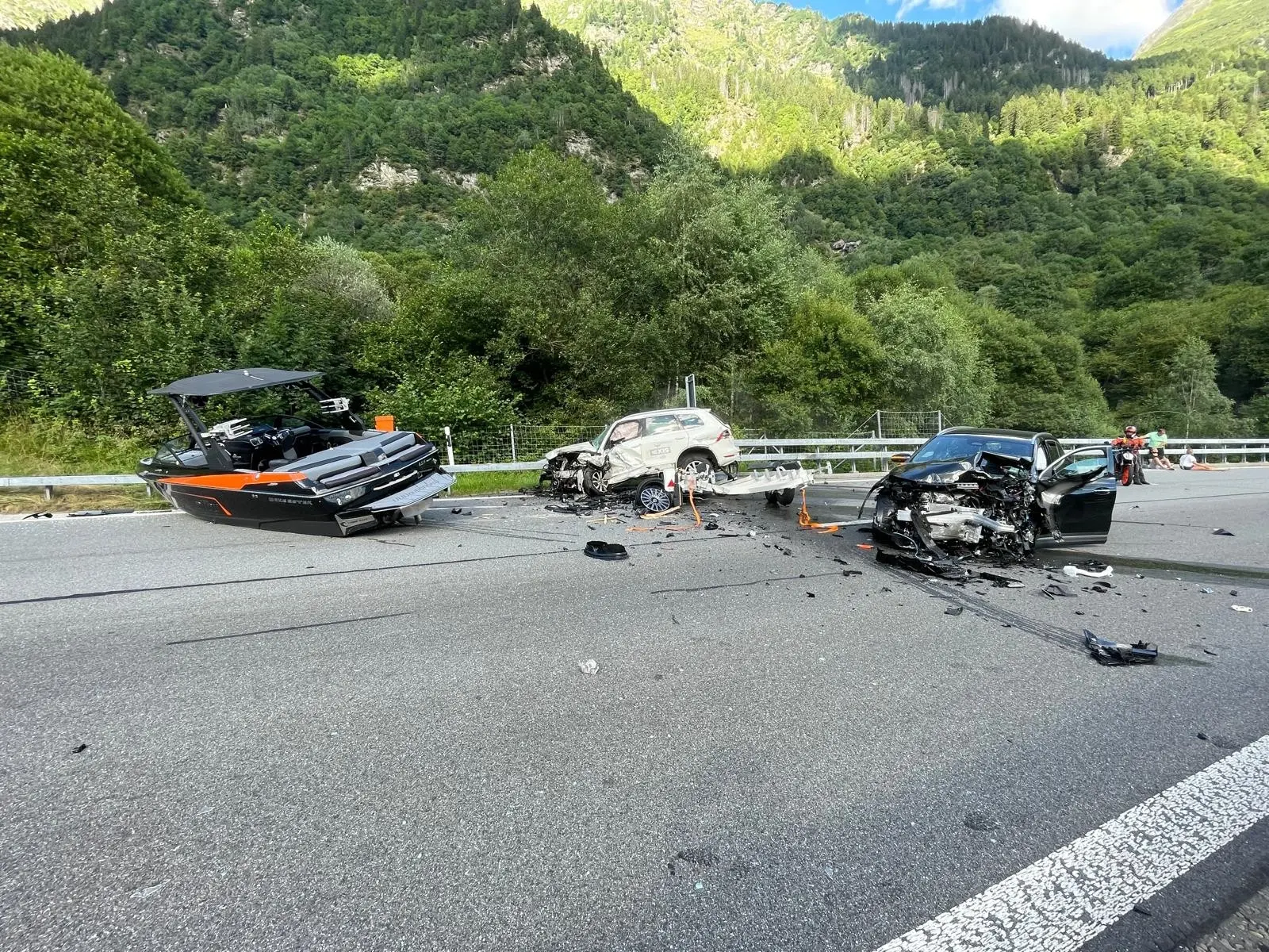 Un grave incidente stradale su una strada di montagna, con un'auto danneggiata e un rimorchio per barche coinvolto. Il paesaggio circostante è verde e montagnoso. I veicoli sono posizionati in modo disordinato, con detriti sparsi sulla carreggiata.