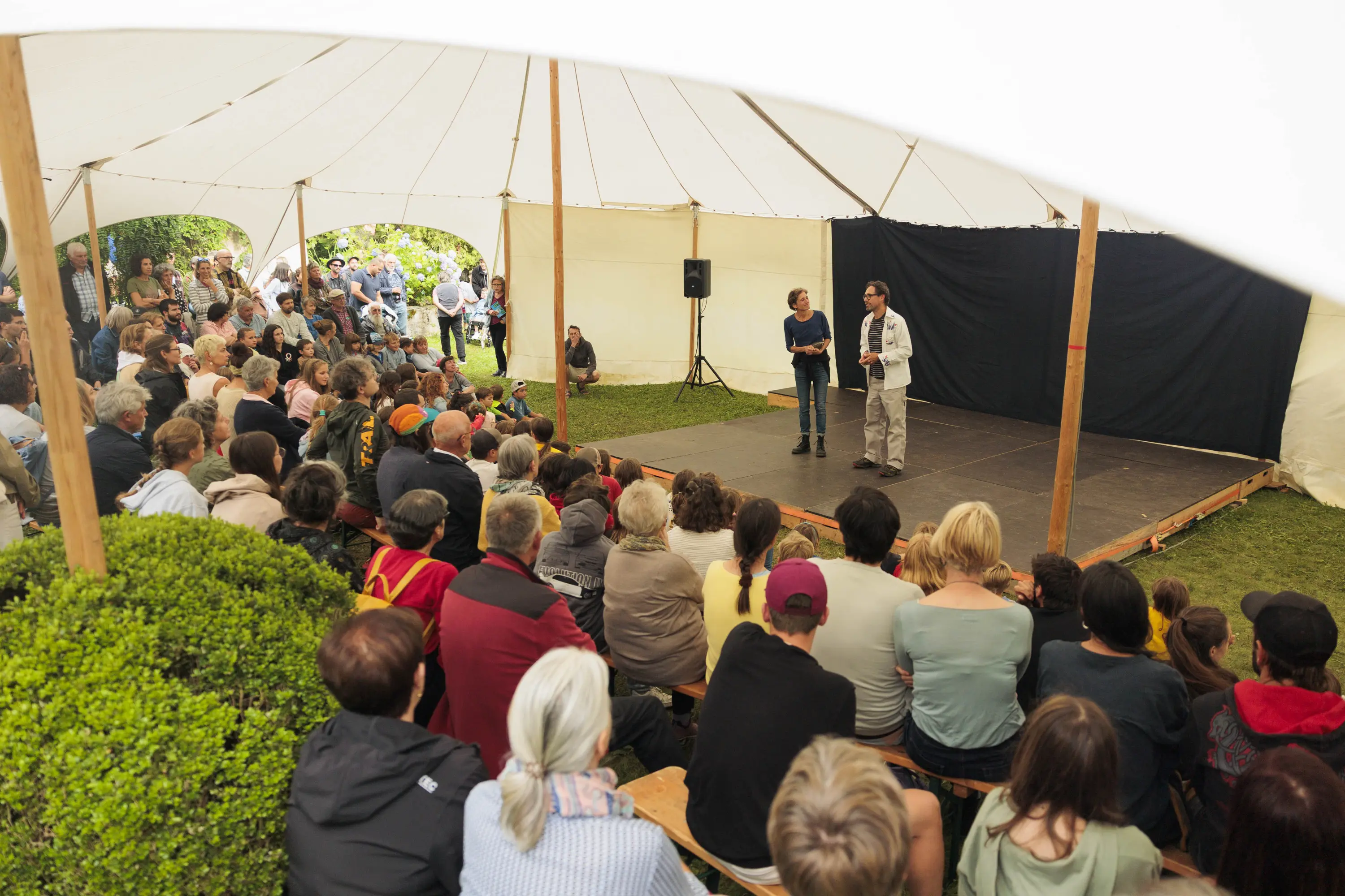 Un gruppo di persone sedute in un tendone, attentamente in ascolto, mentre due relatori si trovano su un palco. Il pubblico sembra coinvolto e curioso.