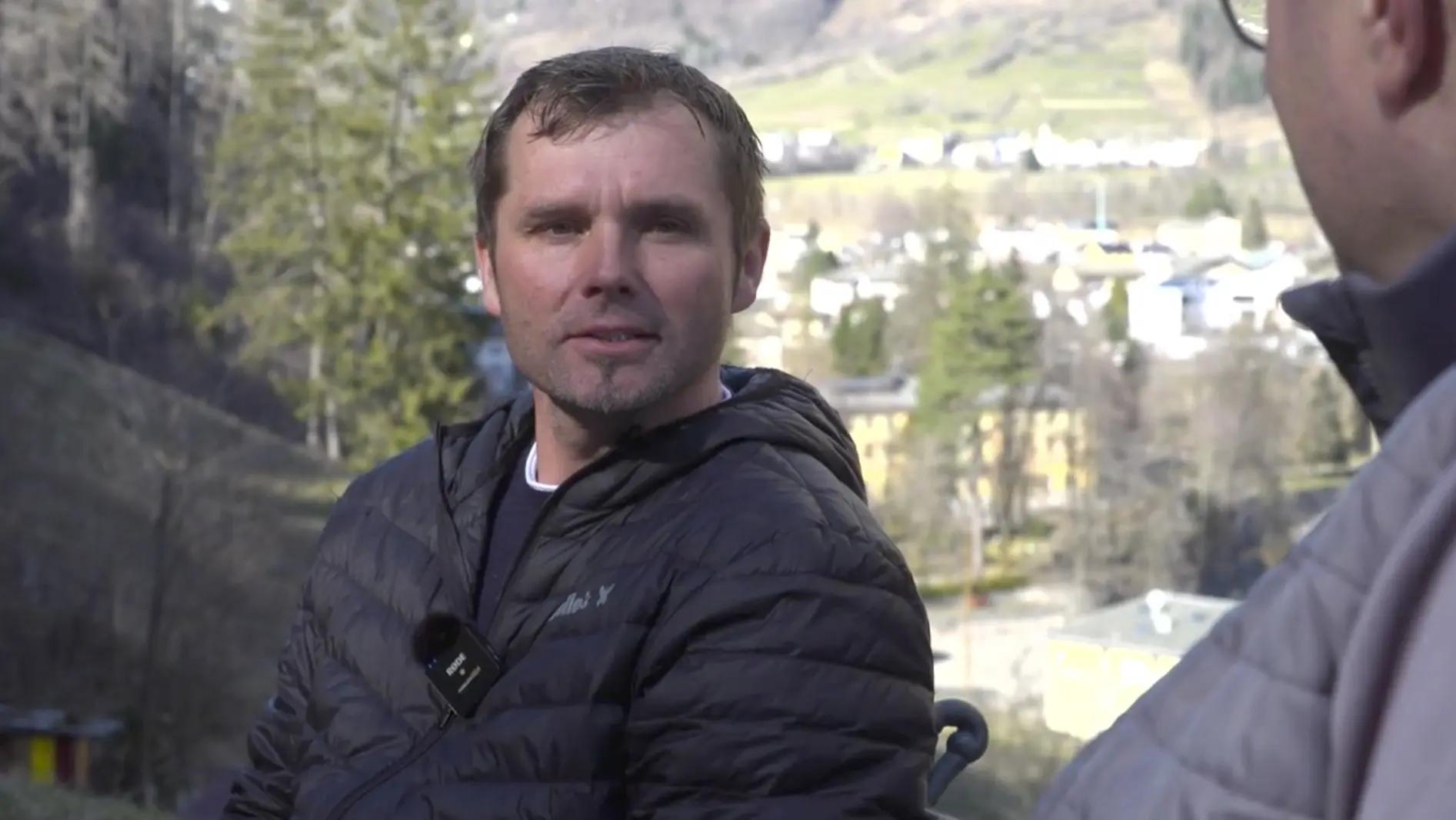 Uomo con giacca nera seduto all'aperto, circondato da un paesaggio montano e verde, con edifici e natura sullo sfondo.