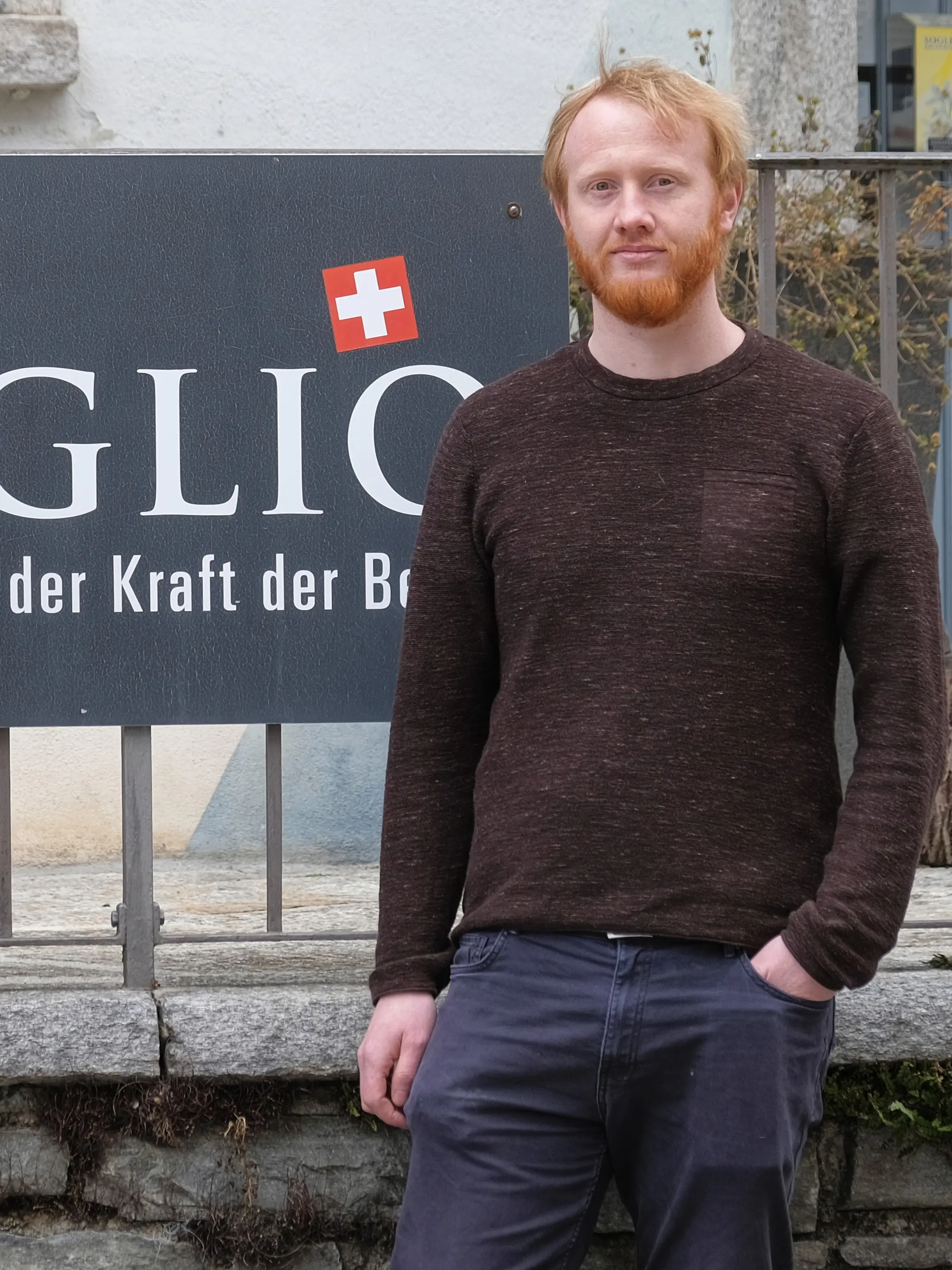 Un uomo con la barba rossa e i capelli biondi arriva in piedi accanto a un cartello che riporta la scritta "GLIO" in un contesto urbano. Indossa un maglione scuro e pantaloni blu.