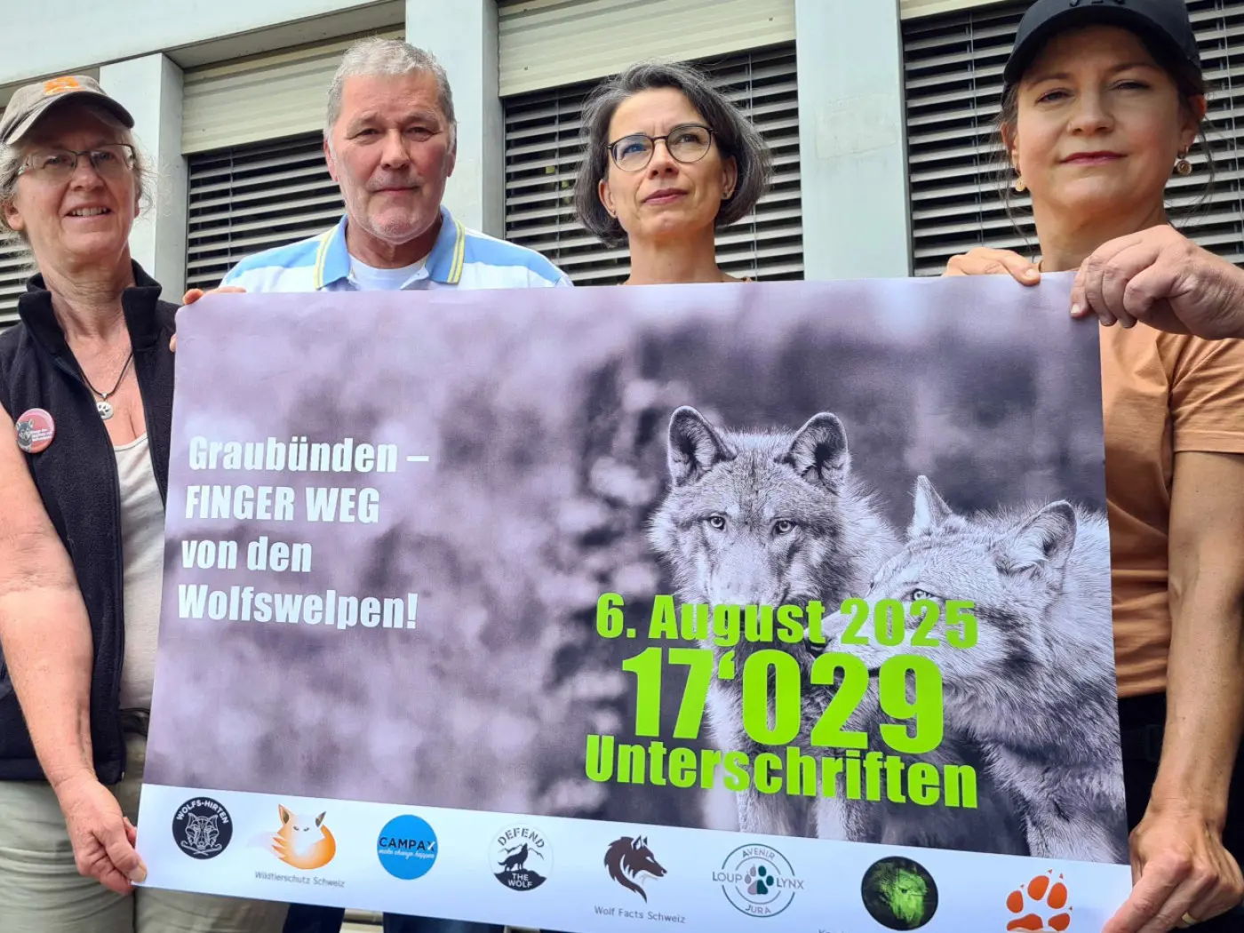 Quattro persone tengono un grande striscione che promuove una campagna per la protezione dei lupi. Lo striscione contiene la scritta "Graubünden - FINGER WEG von den Wolfswelpen!" e la data "6. August 2025" con il numero di firme raccolte, "17'029".