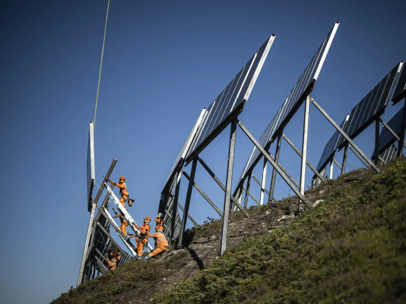 Lavoratori in tute arancioni installano pannelli solari su un pendio. Il cielo è sereno e blu, e si possono vedere i riflessi dei pannelli.