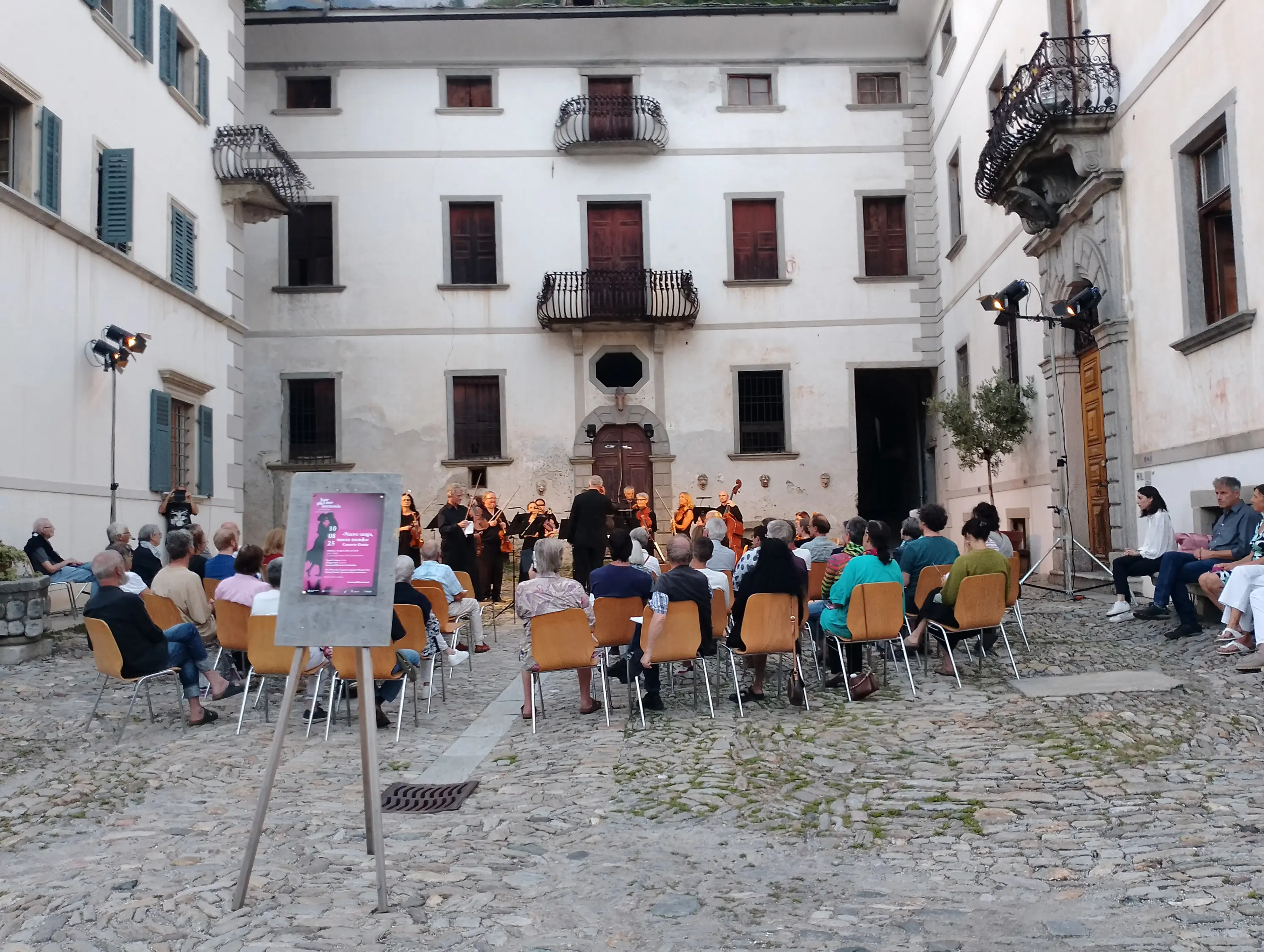 Una platea di persone sedute su sedie di legno, che assistono a una performance musicale in un cortile. Sullo sfondo si vede un edificio storico con finestre e balconi. Un cartello informativo è posizionato in primo piano.