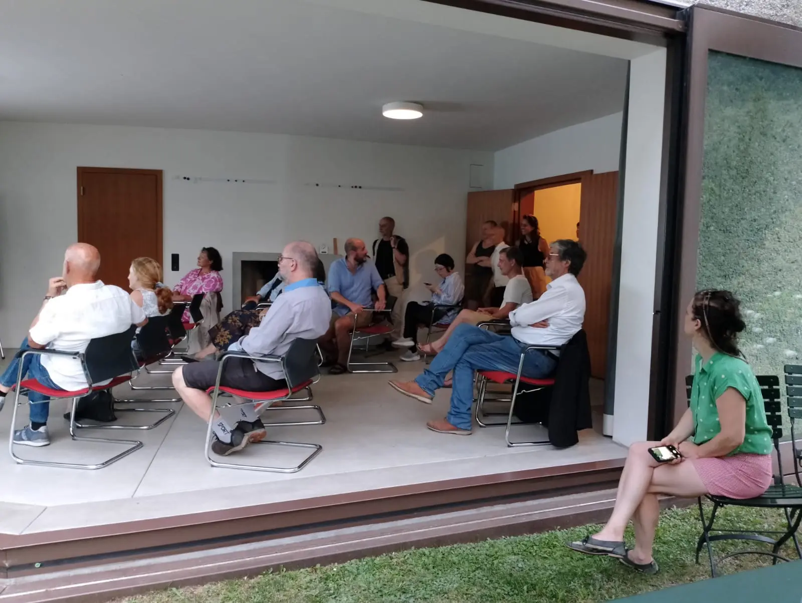 Un gruppo di persone sedute in un ambiente interno con ampie finestre. Alcuni partecipanti sono in piedi, mentre altri ascoltano attentamente. L'atmosfera è serena e informale.