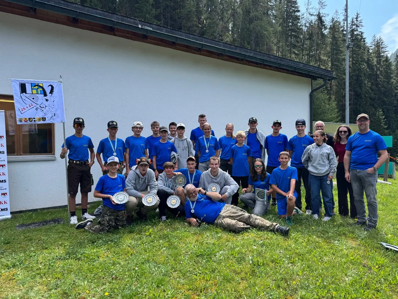 Gruppo di giovani e adulti sorridenti in magliette blu, riuniti all'esterno vicino a un edificio bianco, con trofei e medaglie in evidenza. Sullo sfondo, alberi e una bandiera.
