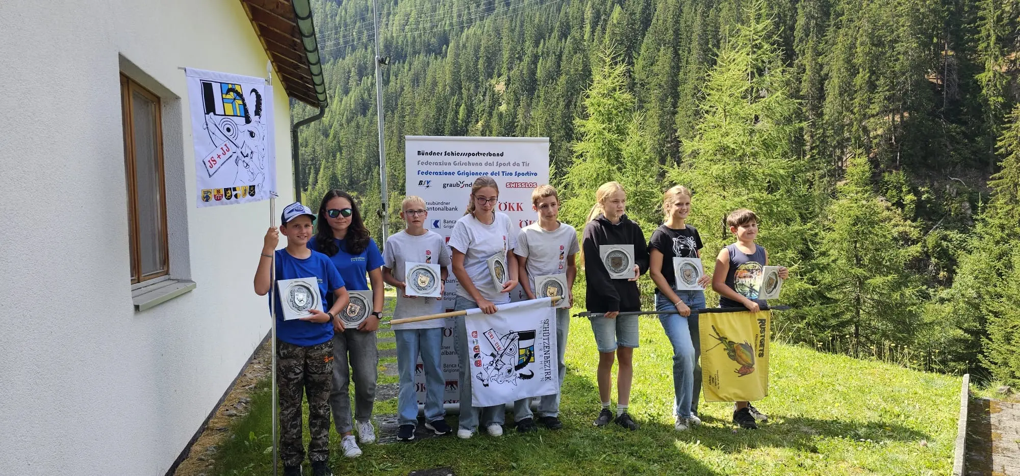 Gruppo di ragazzi sorridenti in un'area verde, alcuni con bandiere e medaglie, davanti a una parete bianca e alberi sullo sfondo.