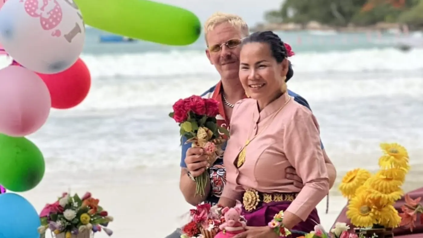 Una coppia sorridente in spiaggia, circondata da decorazioni colorate e fiori. L'uomo tiene in mano un mazzo di fiori, mentre la donna indossa un abito tradizionale e sfoggia un sorriso felice. Sullo sfondo si vedono le onde del mare.