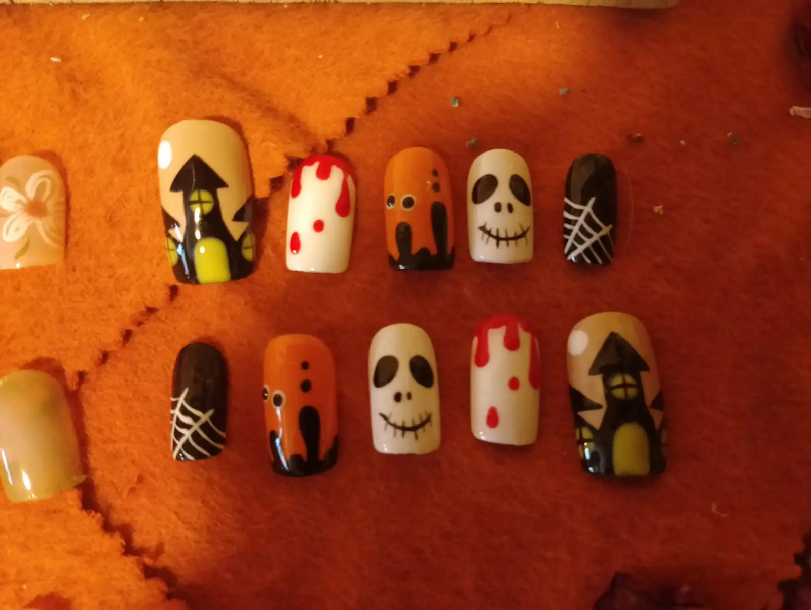 Unghie decorative con design di Halloween. Mostrano teschi, gocce di sangue e ragni su uno sfondo arancione e nero. I colori predominanti sono arancione, bianco e nero.