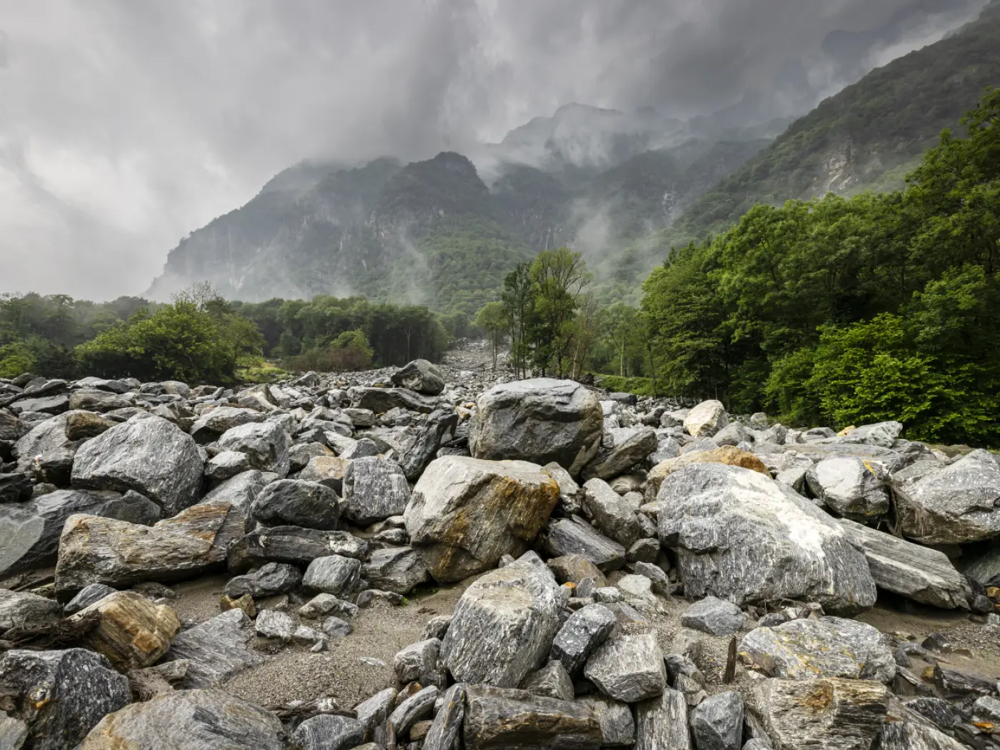 Un paesaggio montano caratterizzato da grandi massi e rocce disposti su un terreno accidentato, circondato da vegetazione verde e alberi. Sullo sfondo, montagne avvolte dalla nebbia e da nuvole scure.