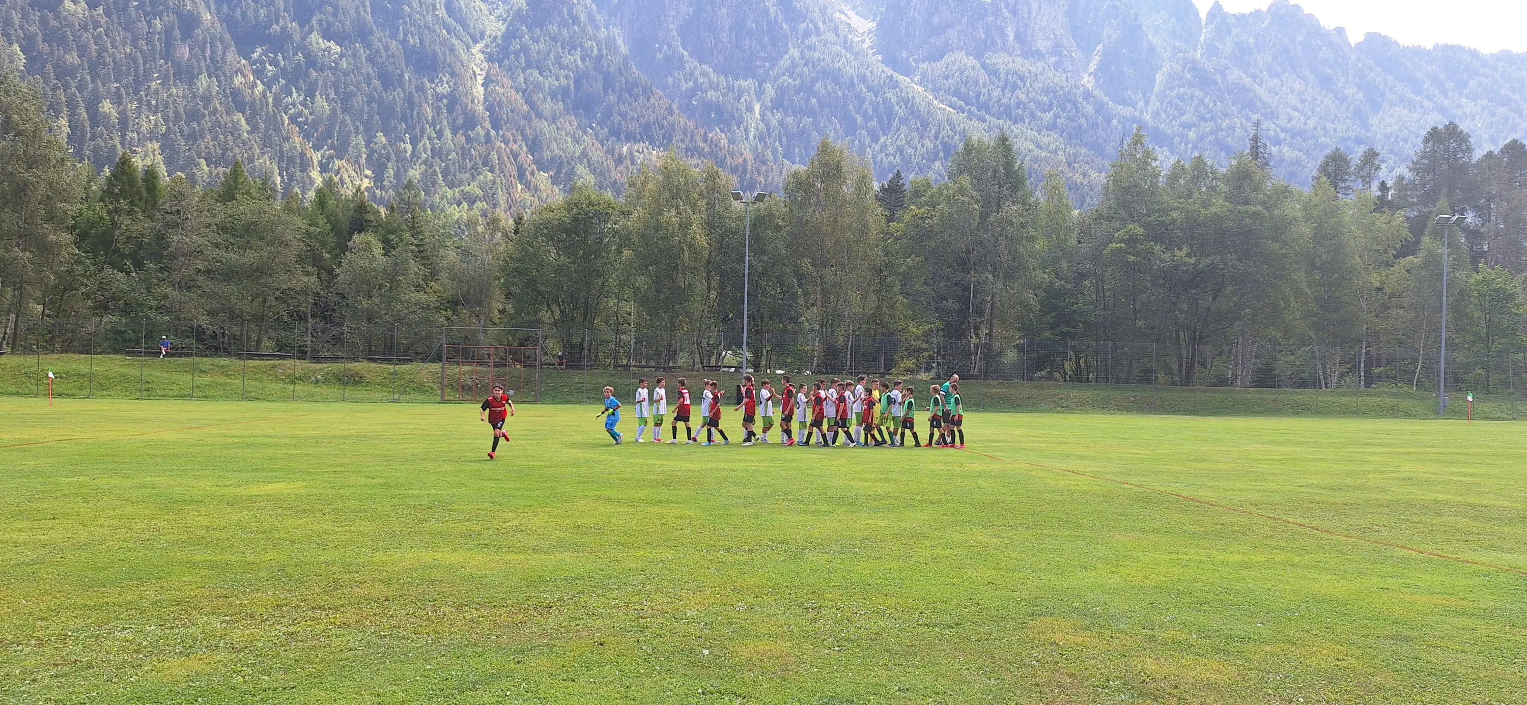 Giocatori di calcio si stringono la mano prima dell'inizio della partita su un campo circondato da alberi e montagne.