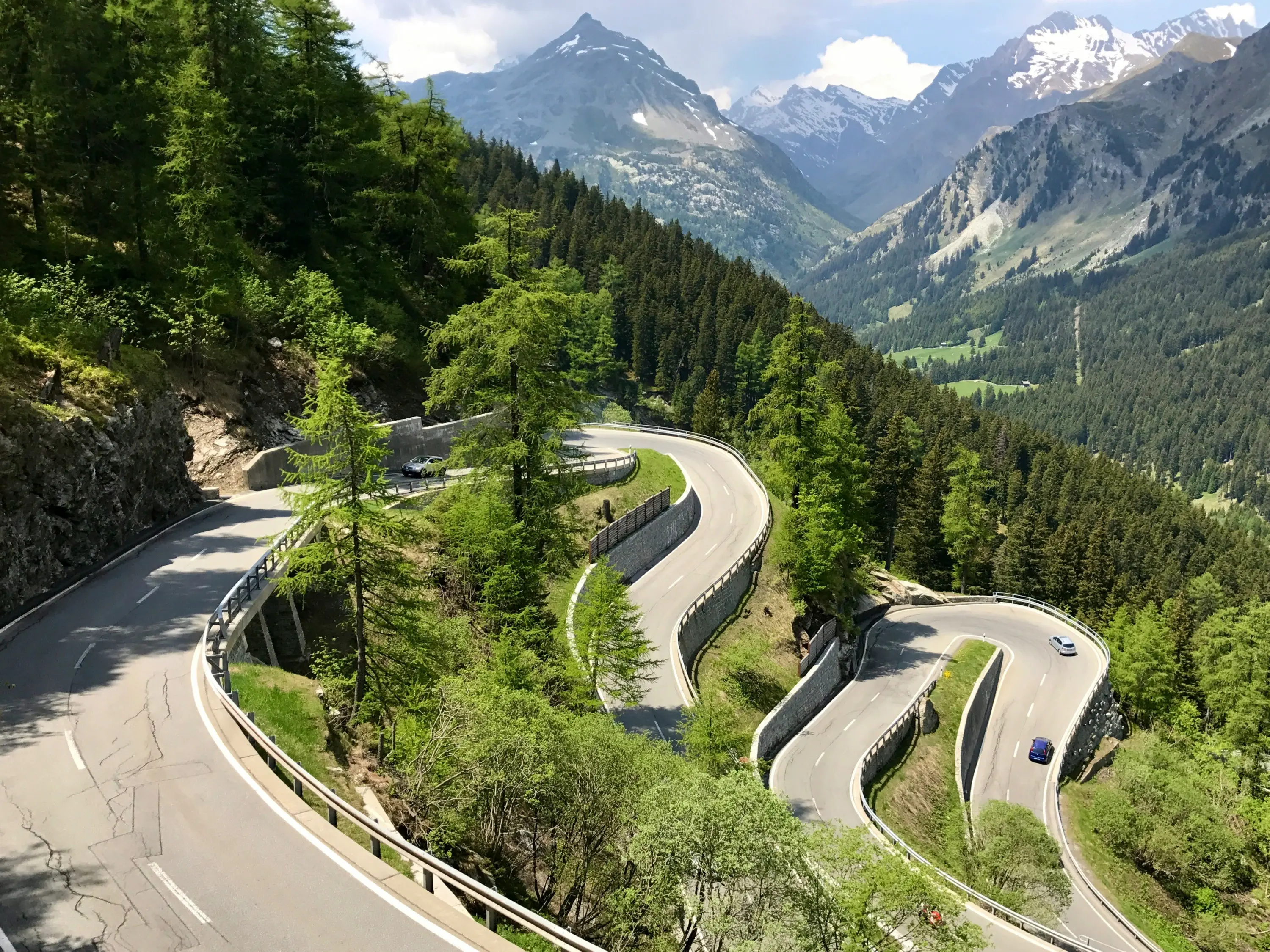 Strada tortuosa che si snoda tra le montagne, circondata da alberi verdi e scoscese colline. Sullo sfondo, vette montuose e un cielo parzialmente nuvoloso.