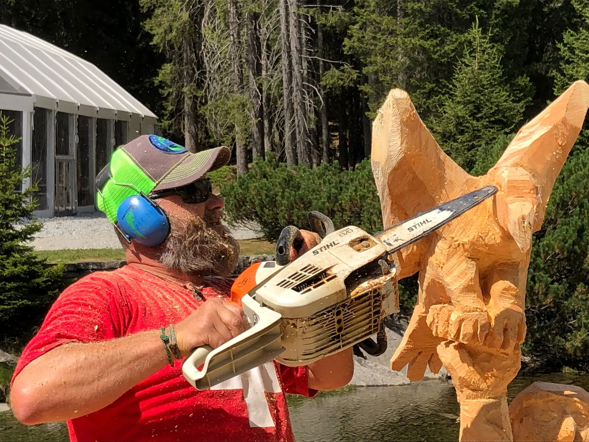 Un uomo con la barba e cuffie protettive sta scolpendo un grande blocco di legno con una motosega, concentrato nel creare una scultura. Sullo sfondo si intravedono alberi e una struttura bianca.